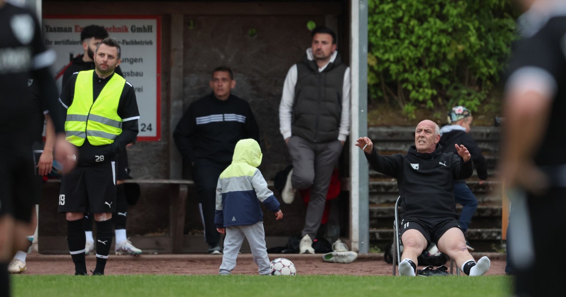 Mario Basler, der rauchend auf einem Klappstuhl am Spielfeldrand sitzt und die Beine von sich streckt