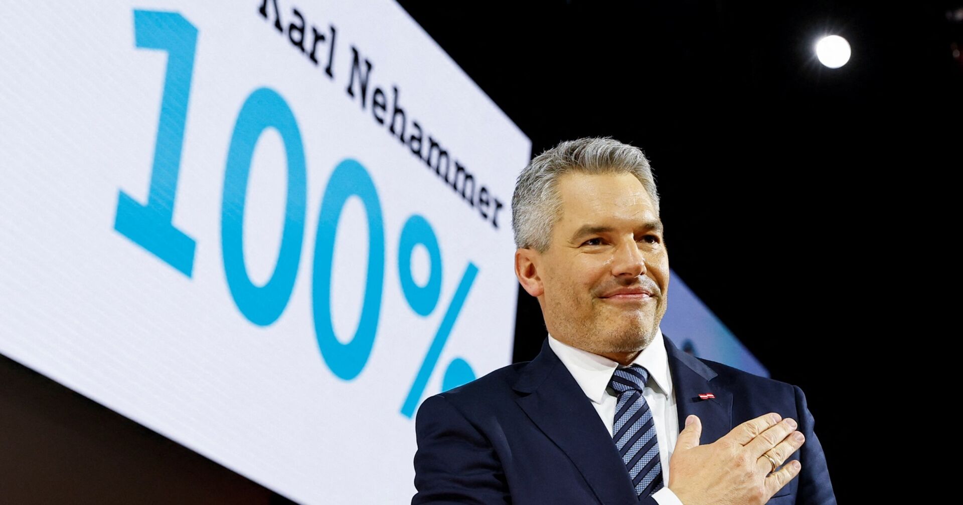 Karl Nehammer vor einem Schild mit der Aufschritt "Karl Nehammer 100 Prozent". Er grinst und hat die Hand am Herz.