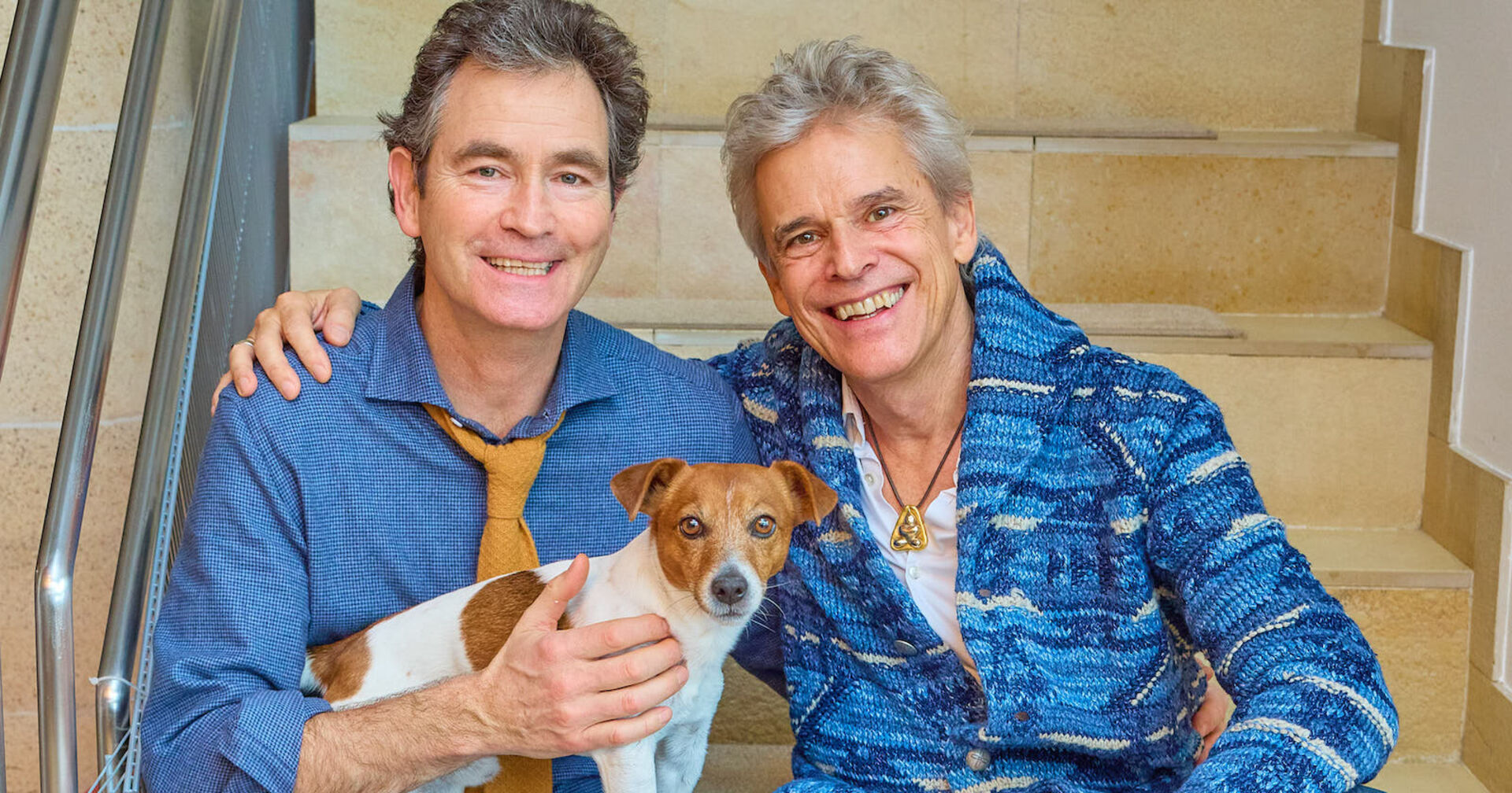 Thomas Brezina und sein Mann sitzen auf Stufen und haben einen Hund am Schoss.
