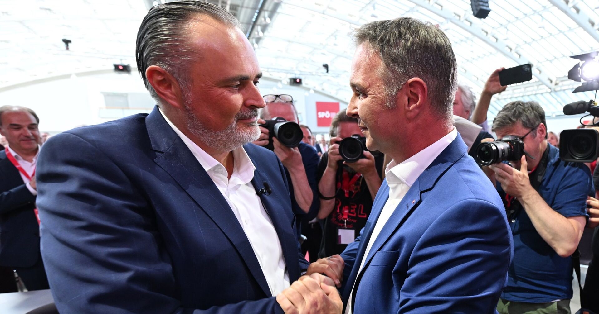 Doskozil und Babler beim Handshake am Parteitag, sie sind von Kameras und Journalisten umgeben.