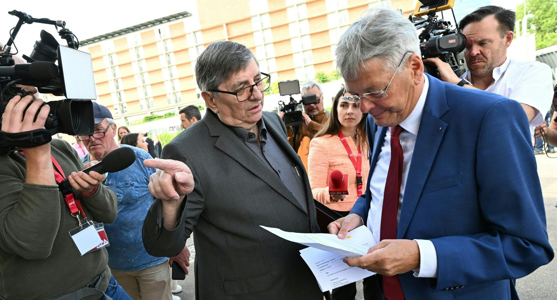 Felber zeigt Kaiser etwas auf einem Zettel und deutet auf den Eingang. Die beiden sind von Kameras umgeben.