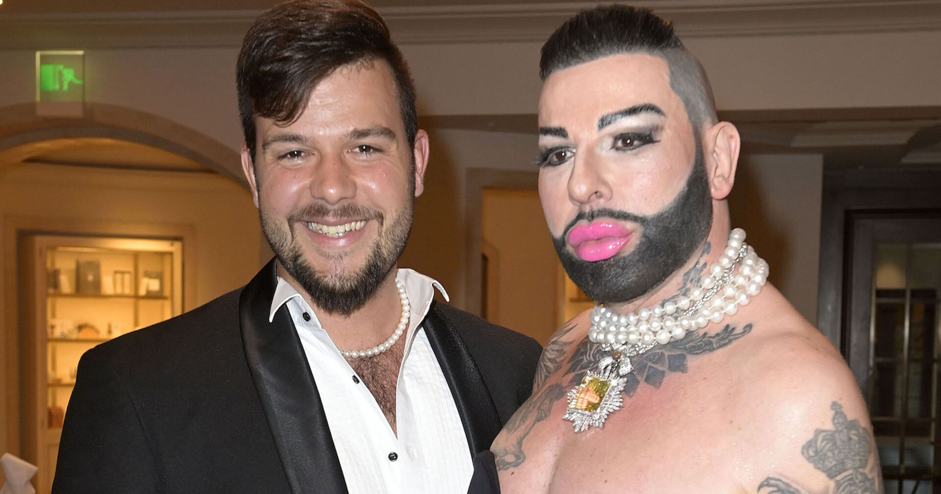 Ein junger Mann mit einer Perlenkette und ein Mann mit Bart und rosa Lippenstift.