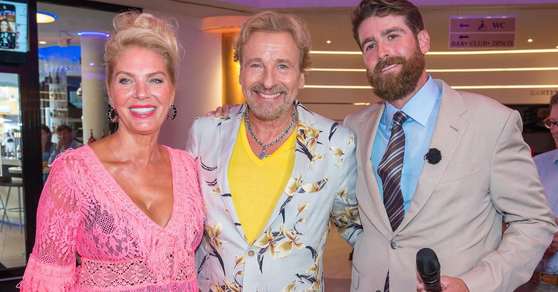 Eine blonde Frau mit einem älteren blonden Mann daneben ein jüngerer Mann mit Bart.