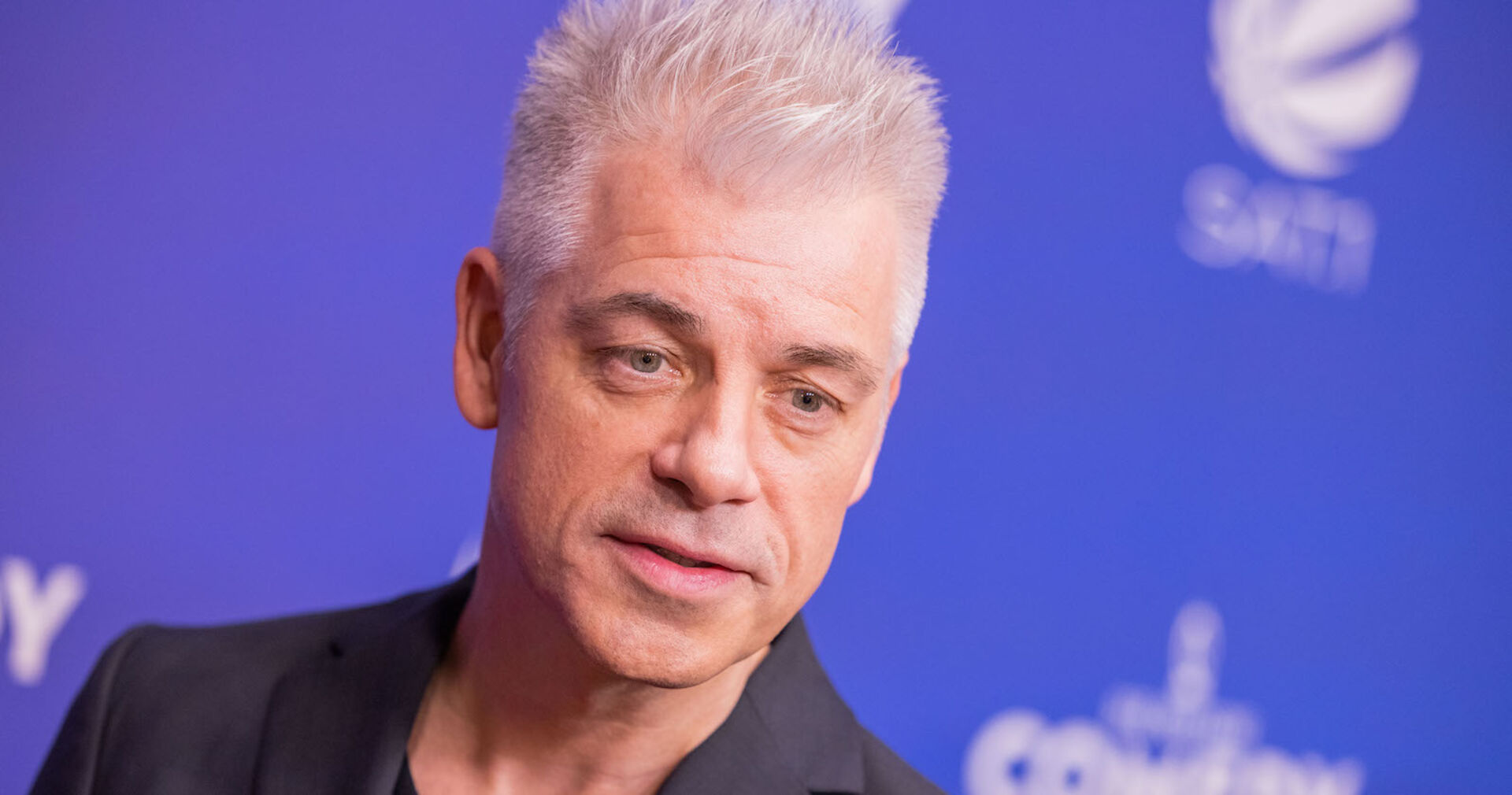 Ein Mann steht vor blauem Hintergrund und hat weiße Haare.