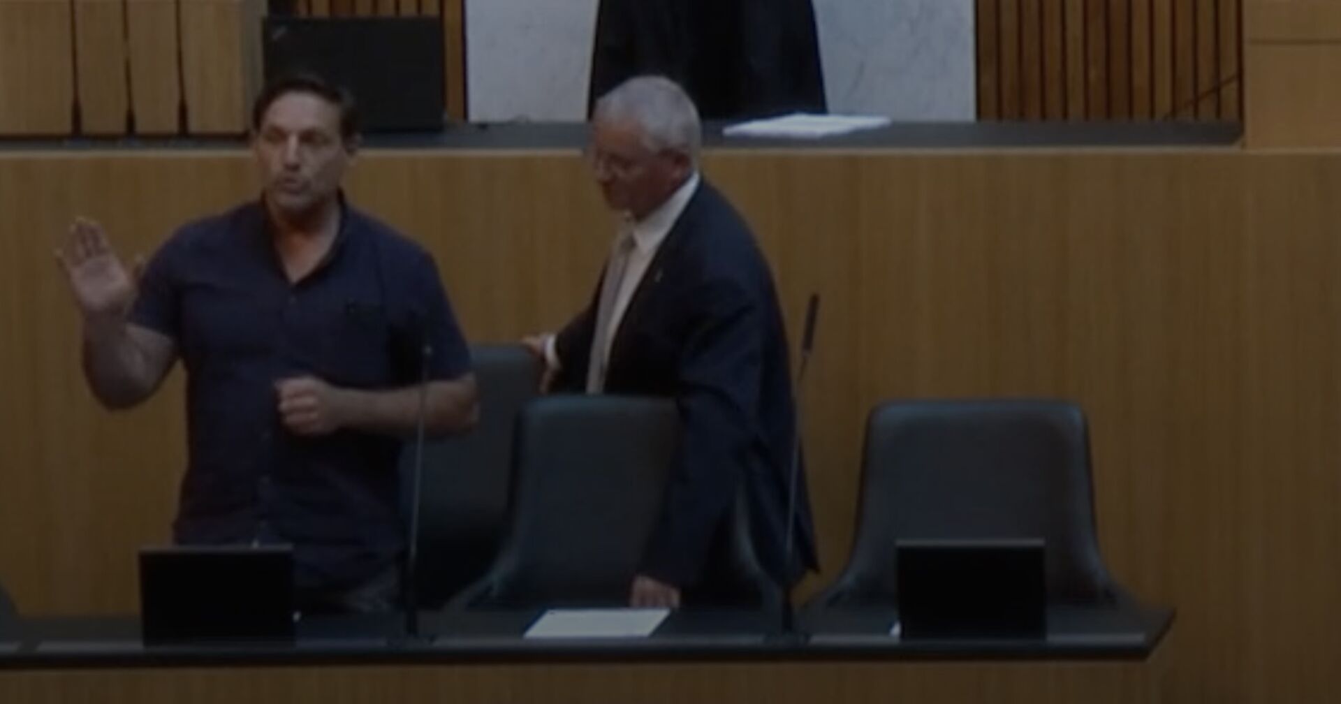 Der Eindringling steht bei der Regierungsbank und redet mit erhobener Hand. Ein Mann in Anzug fasst ihn von der Seite an.