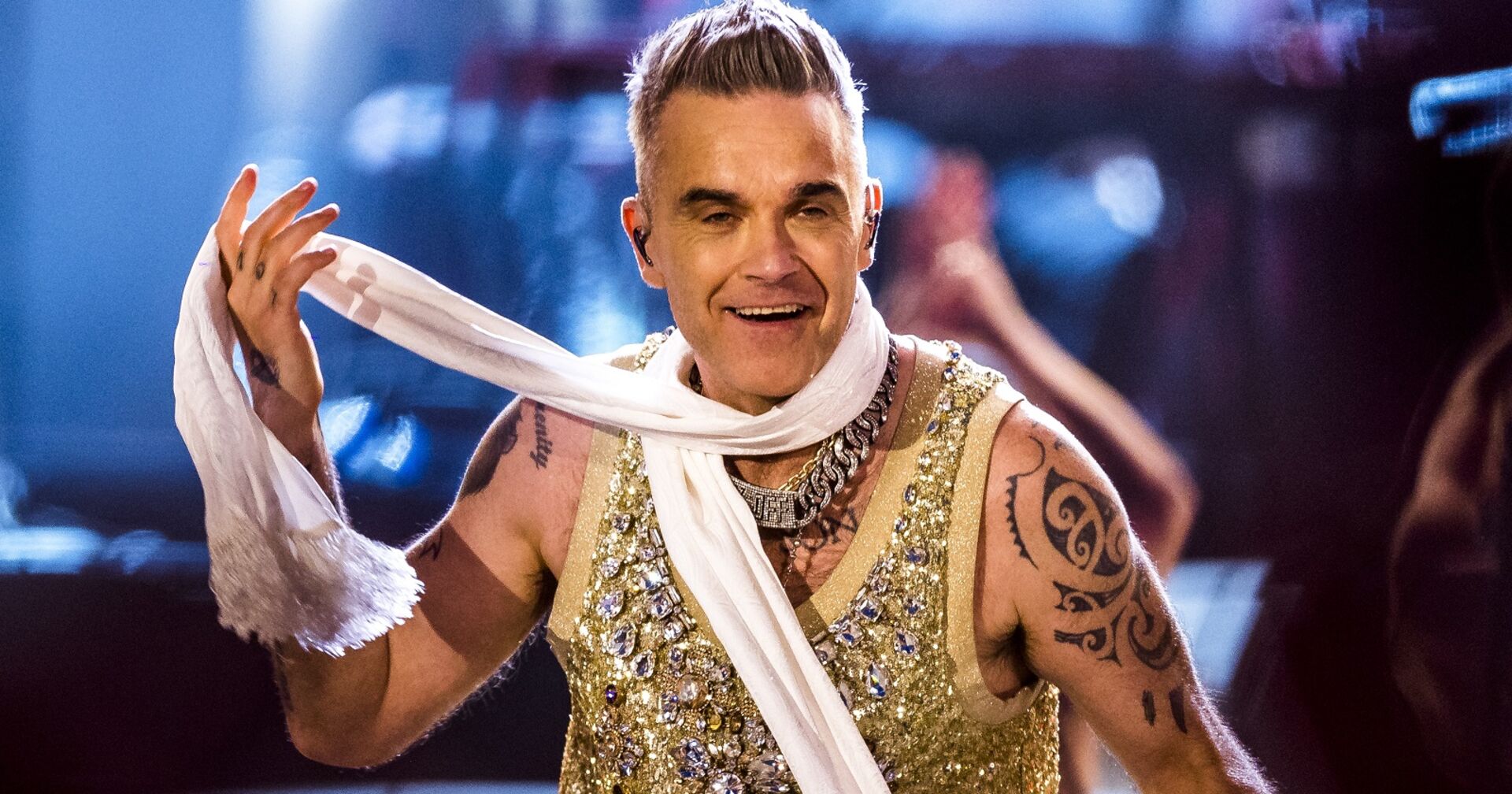 Robbie Williams in einem goldglitzernden Oberteil und einem weißen Schal bei einem Auftritt.