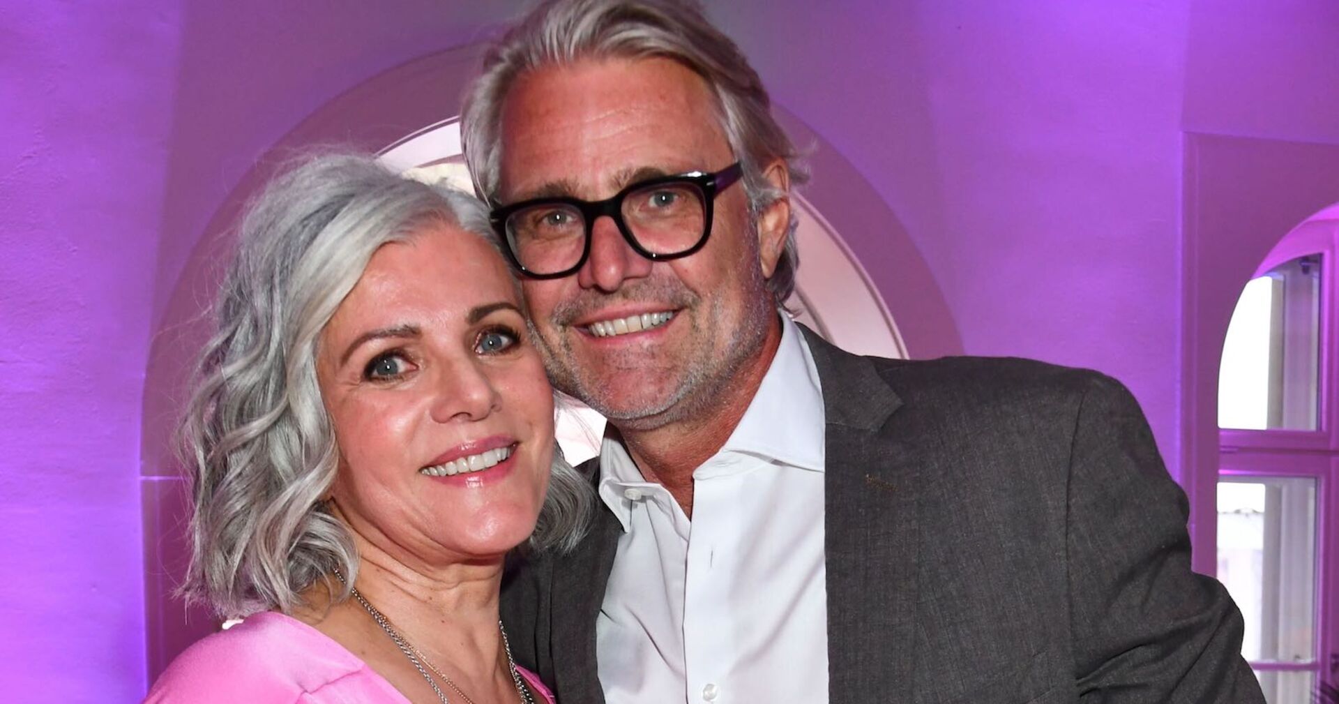 Eine Frau mit grauen Haaren und ein Mann mit einer schwarzen Brille lachen in die Kamera.