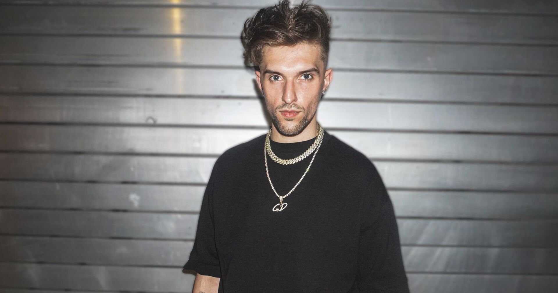 Gavis Dean, der ein schwarzes T-Shirt und eine Goldkette trägt, und vor einem Rolltor steht