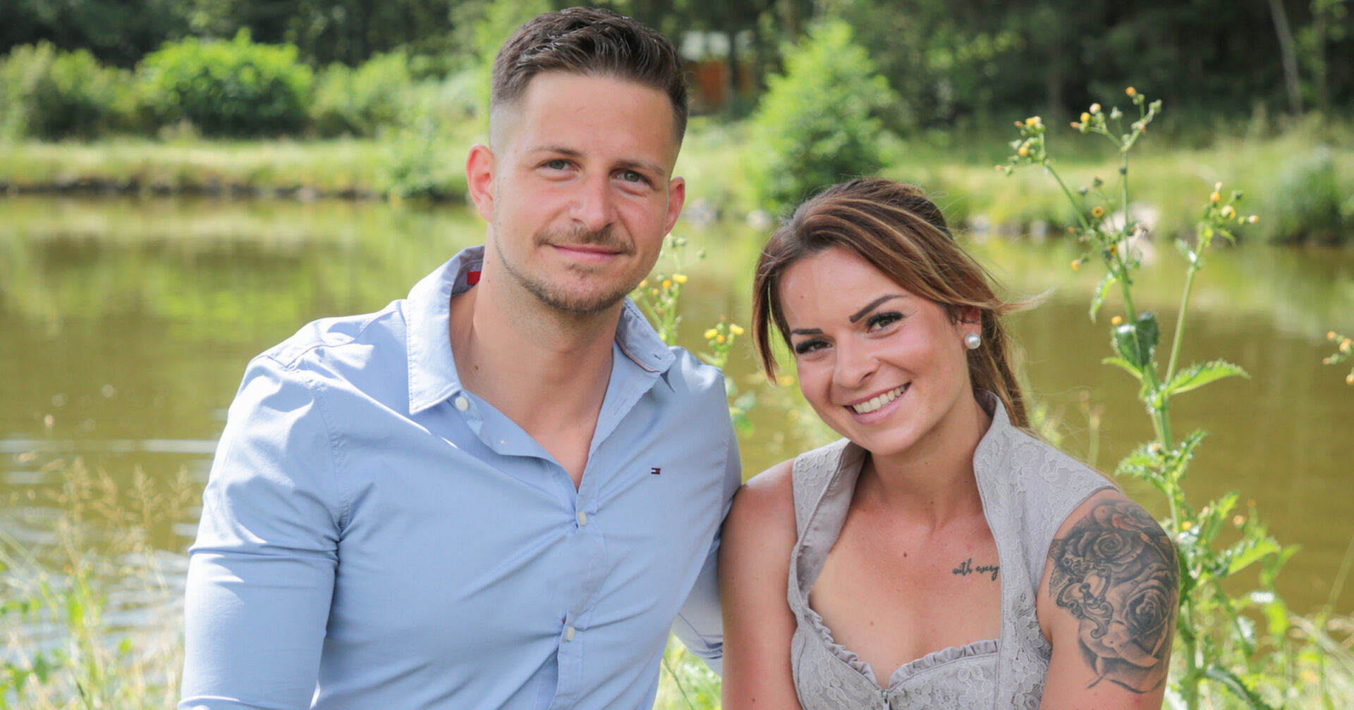 Stefan mit einem hellblauen Hemd und Nicole mit einer Tätowierung am Arm sitzen vor einem See.