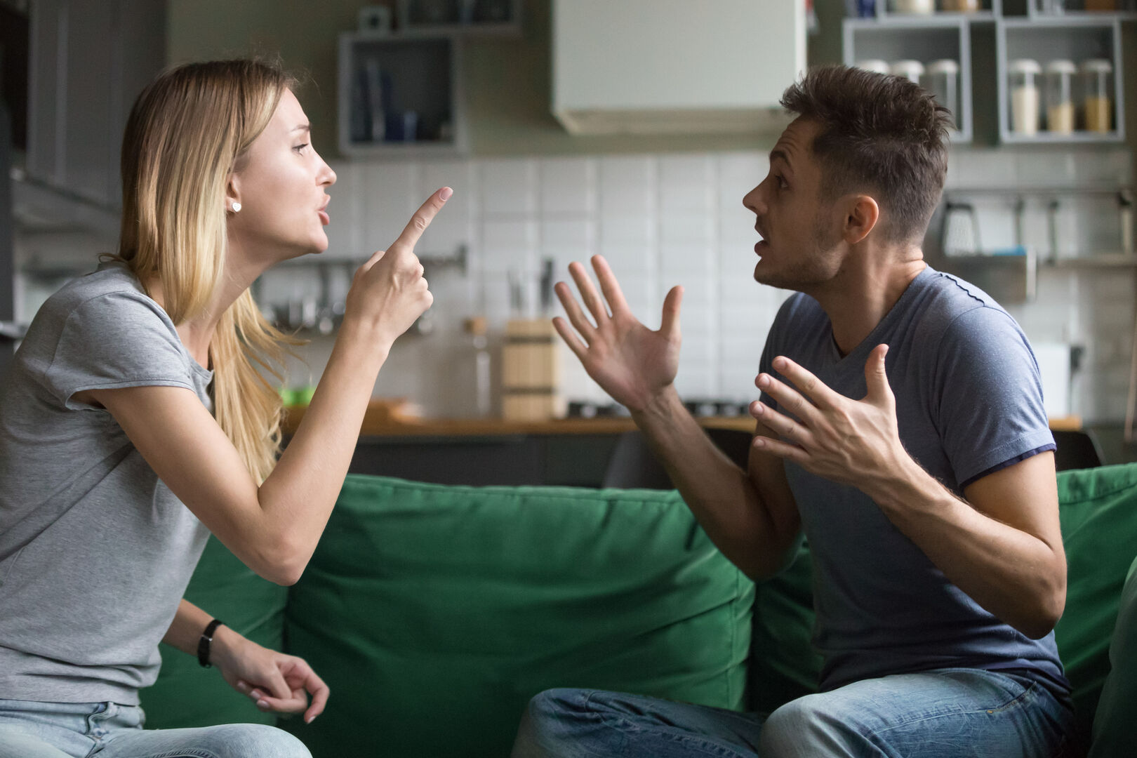 Blonde Frau streitet mit ihrem Mann auf einer grünen Couch vor einer Küche.