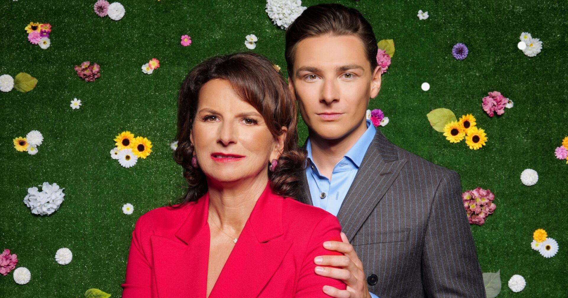 Claudia Obert in einer roten Bluse mit ihrem Partner Max vor einer grünen Wand mit bunten Blumen.