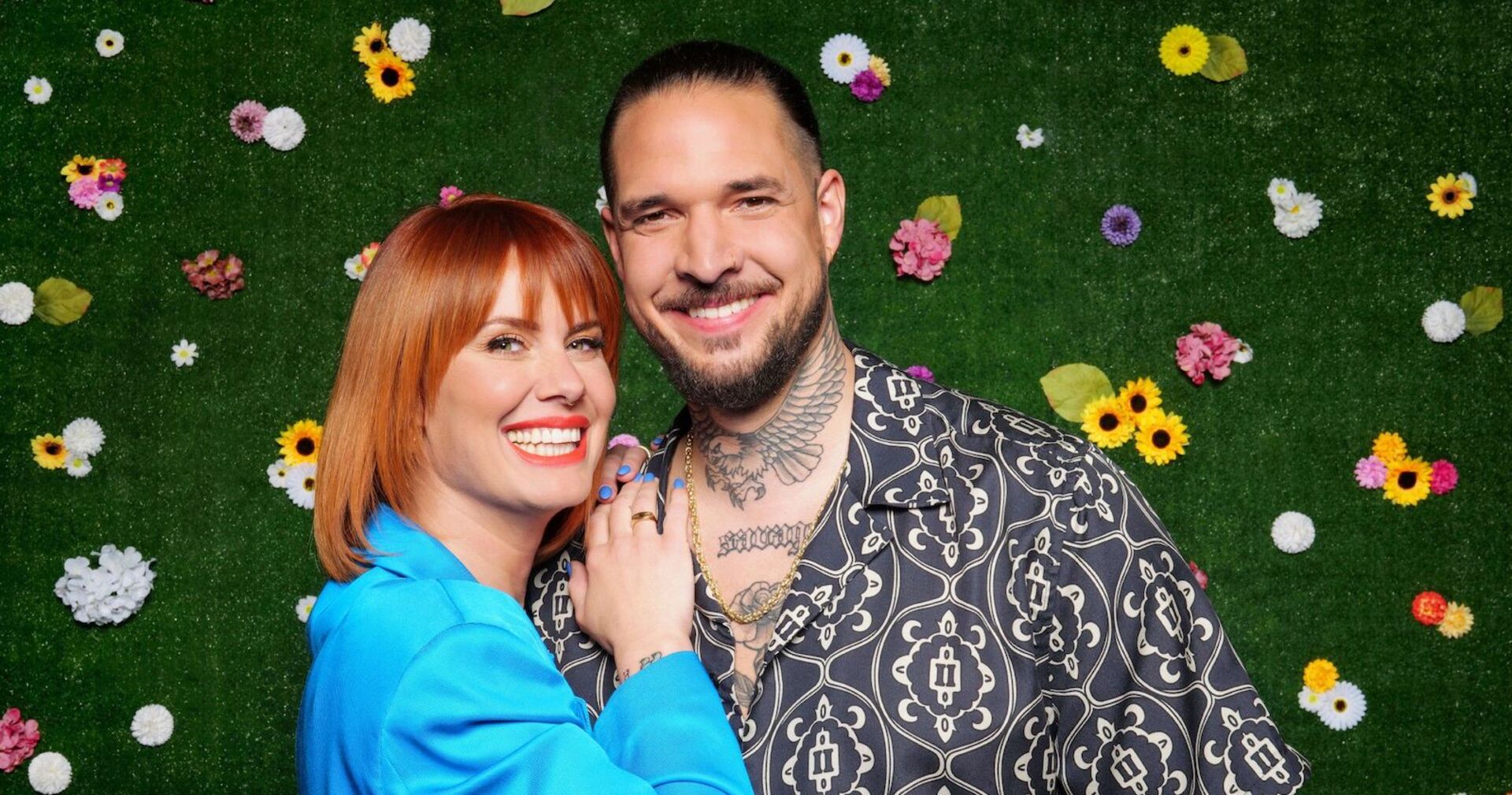 Eine Frau mit schulterlangen Kupfer Haaren und einem blauen Blazer neben einem Mann mit Tattoos. Beide stehen vor einer Wand mit Blumen