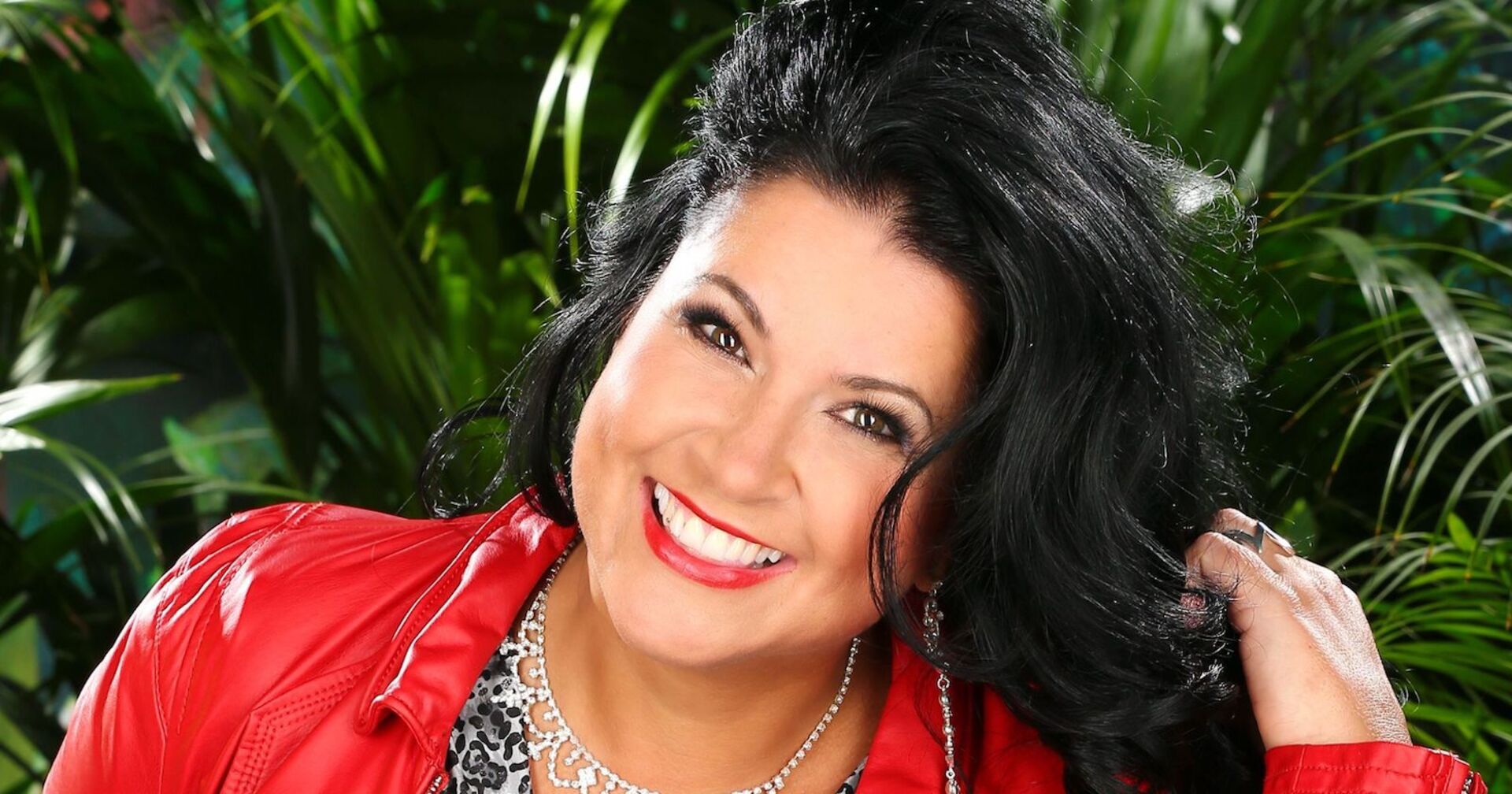 Eine Frau mit einer roten Lederjacke und schwwarzen Haaren. Sie steht vor vielen grünen Palmen.