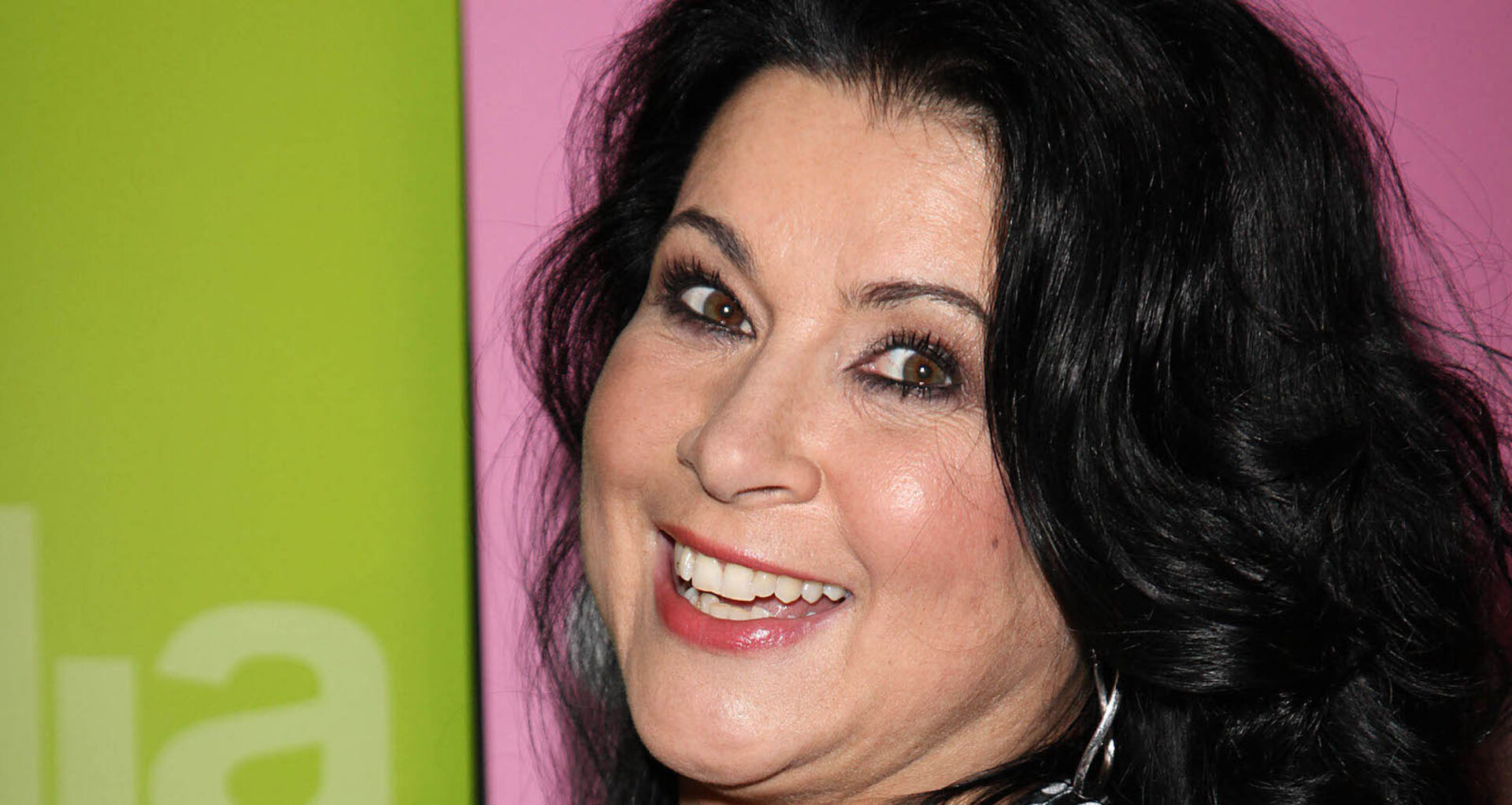 Iris klein lacht in die Kamera mit schwarzen Haaren vor einer grün pinken Wand