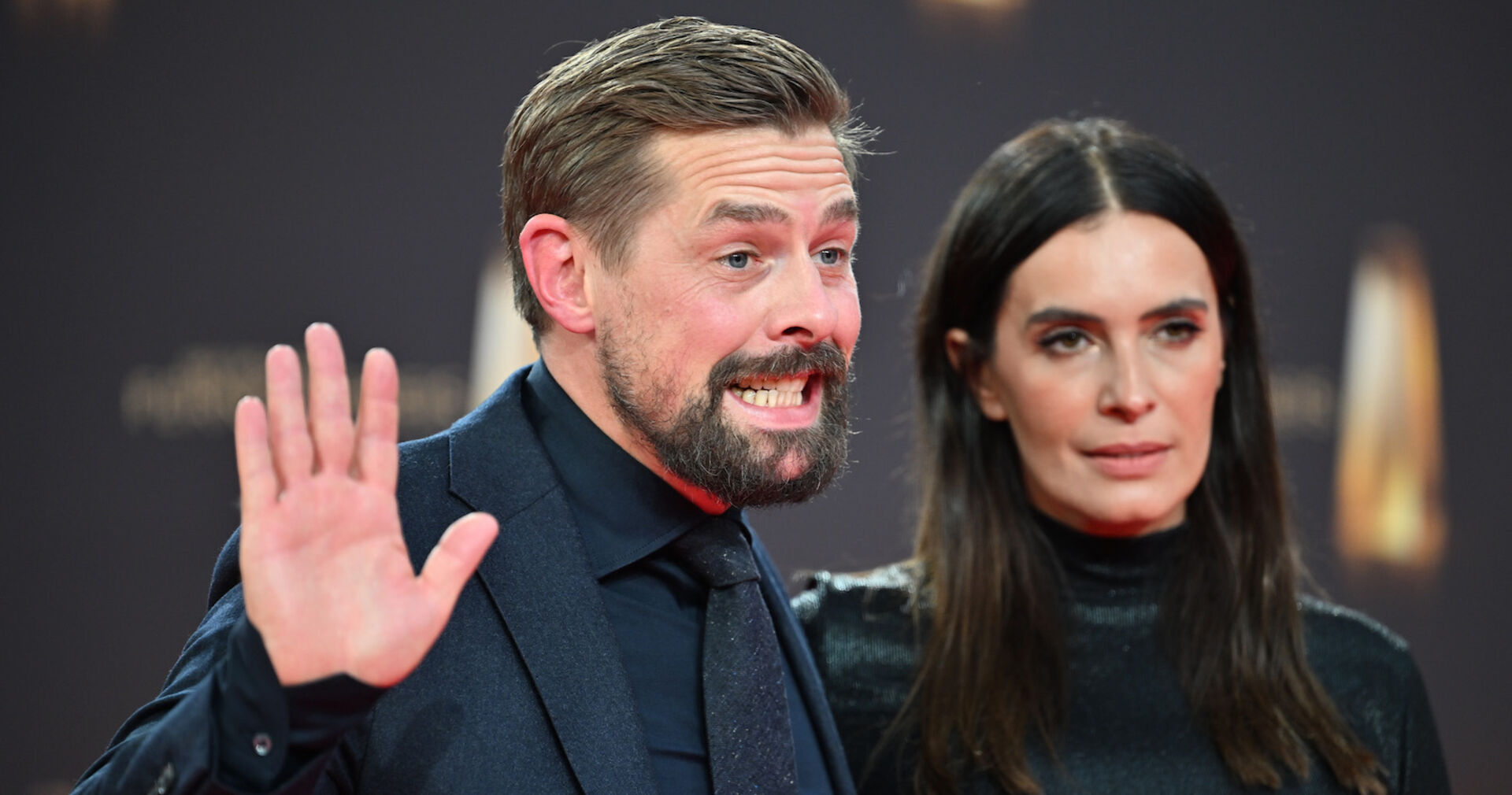 Ein Mann mit kurzen braunen Haare und Vollbart winkt den Fotografen zu. Neben ihm eine Frau mit brünetten offenen Haaren und einem hochgeschlossenen schwarzen Kleid.