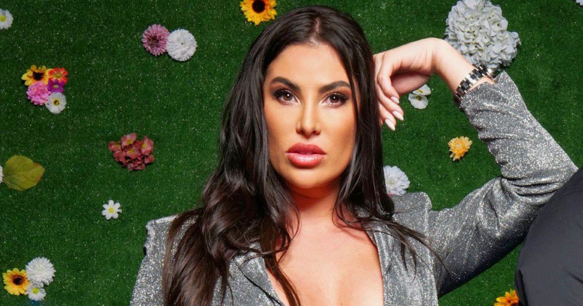 "Sommerhaus der Stars"-Teilnehmerin Ricarda Raatz in einem silbernen Kleid mit schwarzen Haaren vor einer grünen Wand mit Blumen.