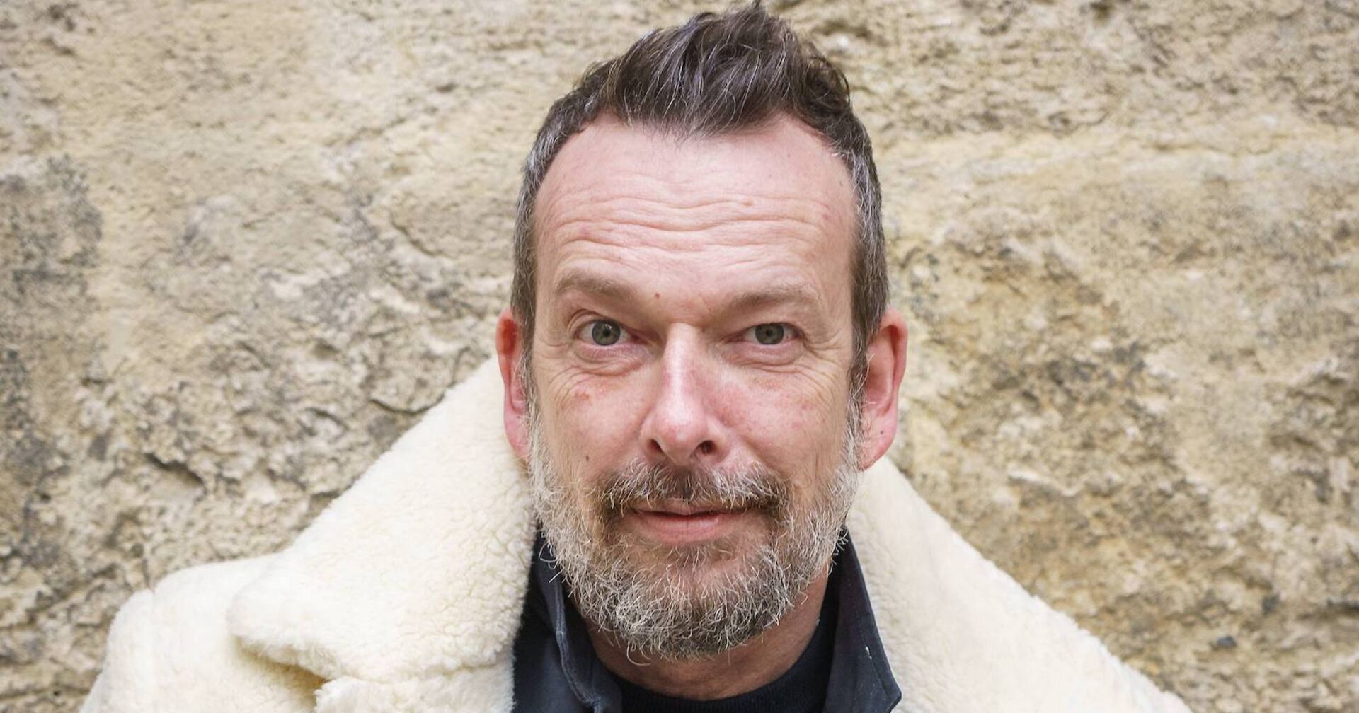 Robert Kratky mit kurzen aufgestellten Haaren in einem weißen Mantel vor einer Mauer.