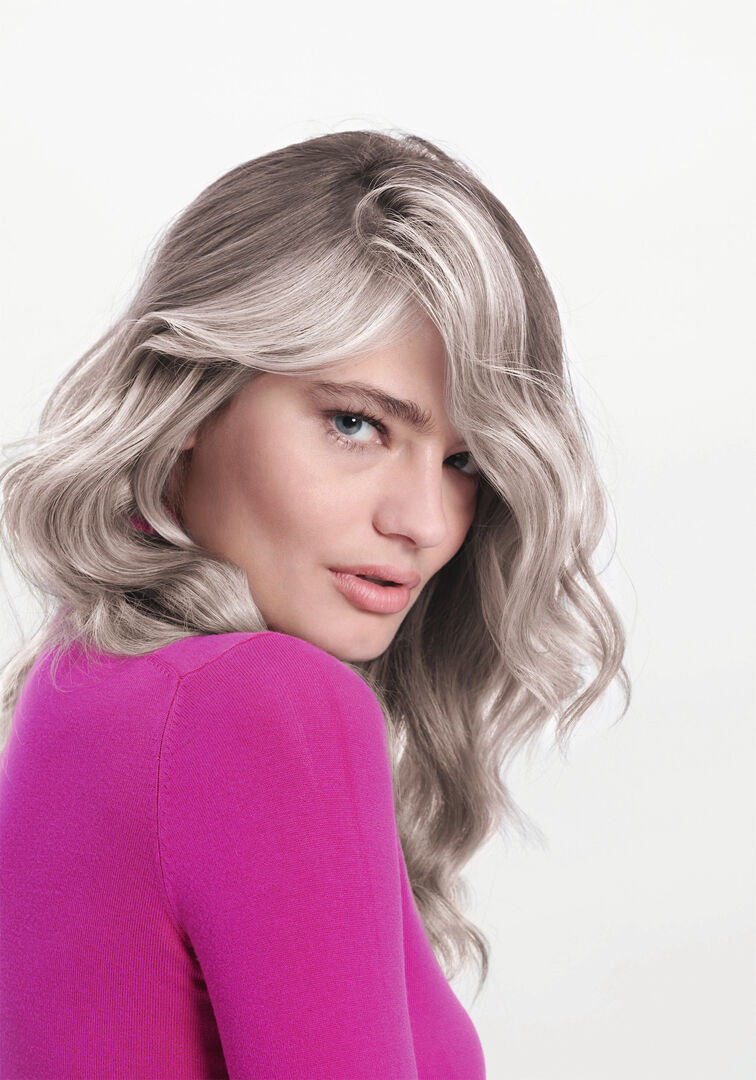 Frau mit blonden Haaren und weißen Strähnen vor einer weißen Fotowand mit einem pinken Pullover.