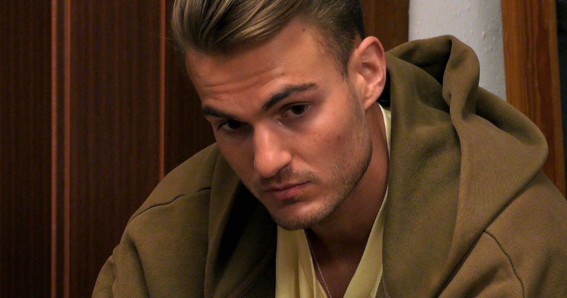 Can ist ein Teilnehmer im Sommerhaus der Stars und sitzt nachdenklich. Er trägt einen olivgrünen Hoodie.