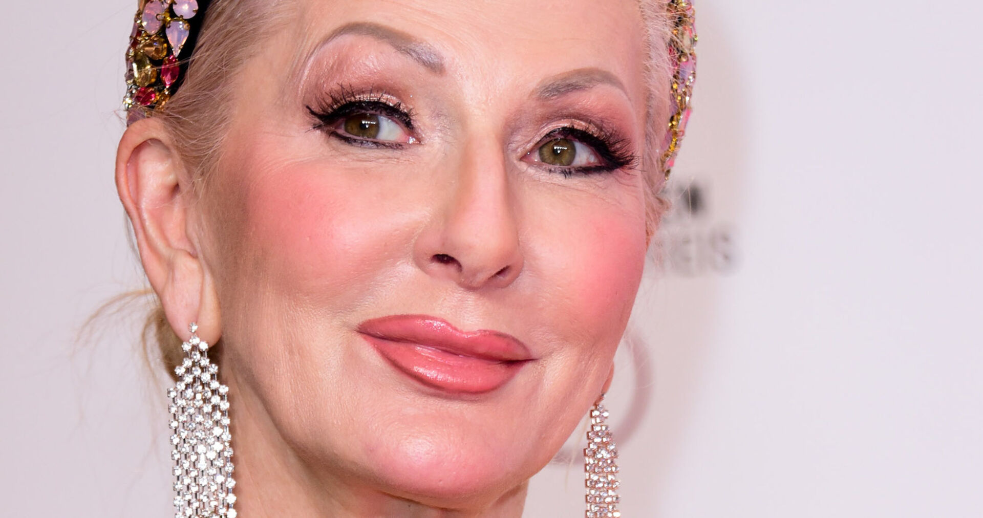 Désirée Nick mit einem starken Make Up vor einer Leinwand mit geschlossenen Haaren und großen silber Ohrringen.