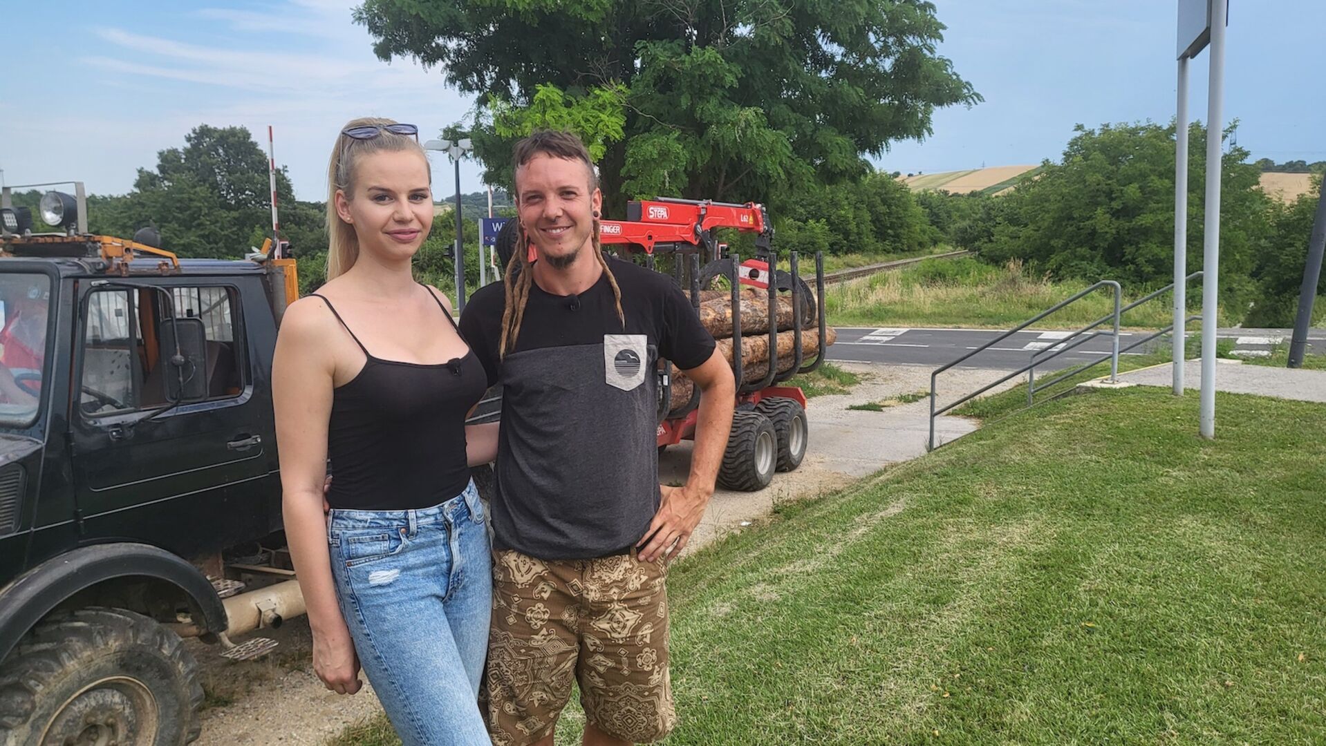 Daniella von Bauer sucht Frau trägt hochgebundene blonde Haare und ein schwarzes Tanktop. Bauer Philip ein schwarzes T-Shirt. Beide stehen vor einem Anhänger.