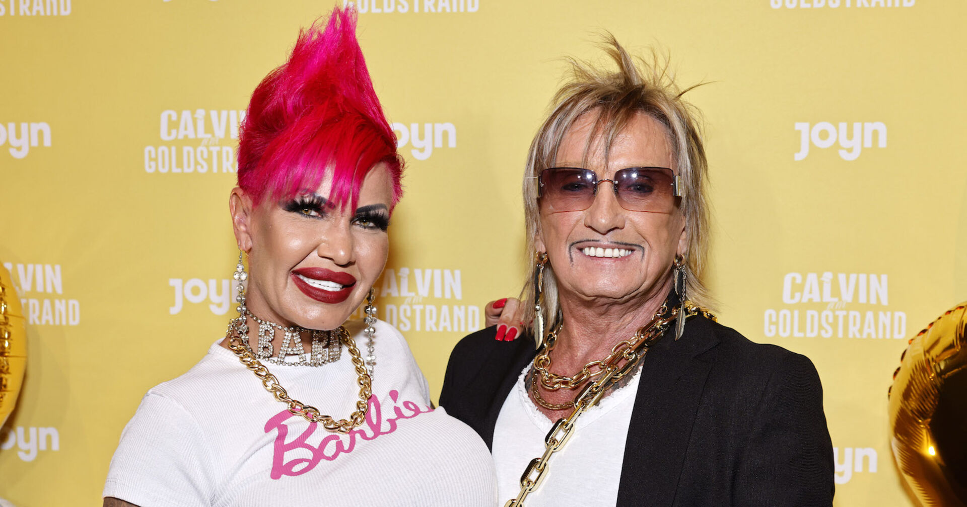 Gina trägt einen pinken Irokesen und ein weißes T-Shirt mit einer Aufschrift "Barbie". Bert Wollersheim hat blonde Haare und eine verdunkelte Sonnenbrille.