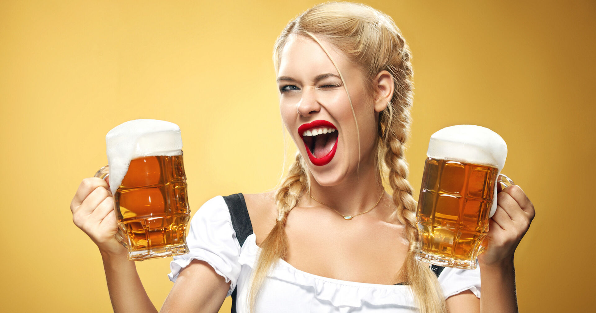 Eine blonde Frau im Dirndl mit zwei geflochtenen Zöpfen hält zwei Bierkrüge und zwinkert in die Kamera.
