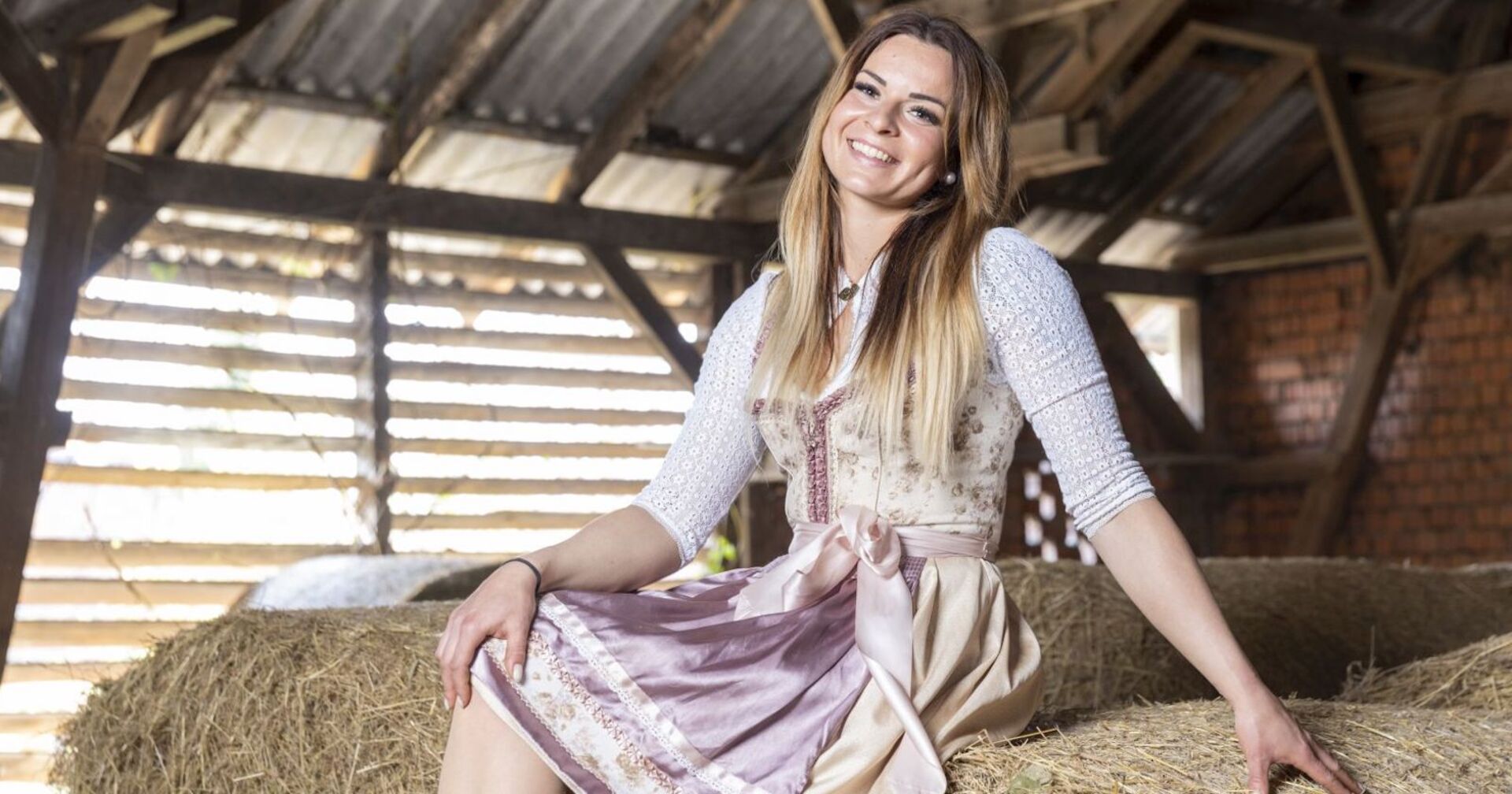 Kandidatin Nicole von Bauer sucht Frau posiert auf Strohballen mit einem rosa Dirndl und offenen blonden Haaren.