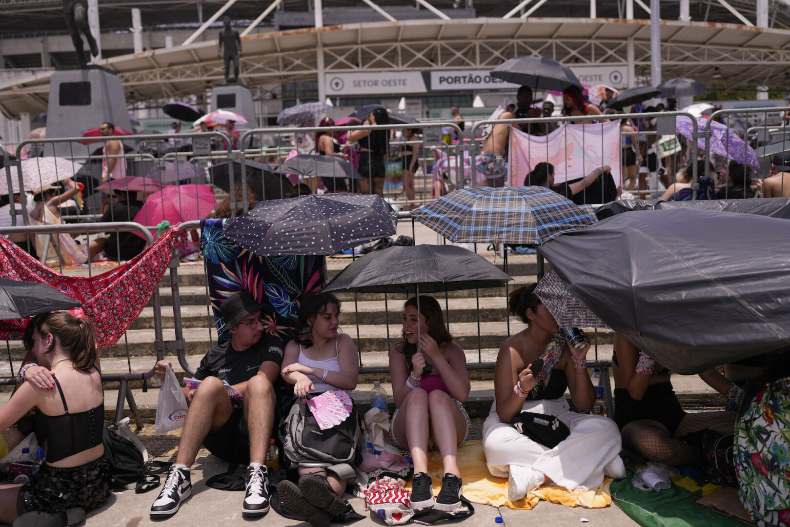 Fans sitzen in Brasilien vor einer großen Konzerthalle und suchen Schutz unter Schirmen.