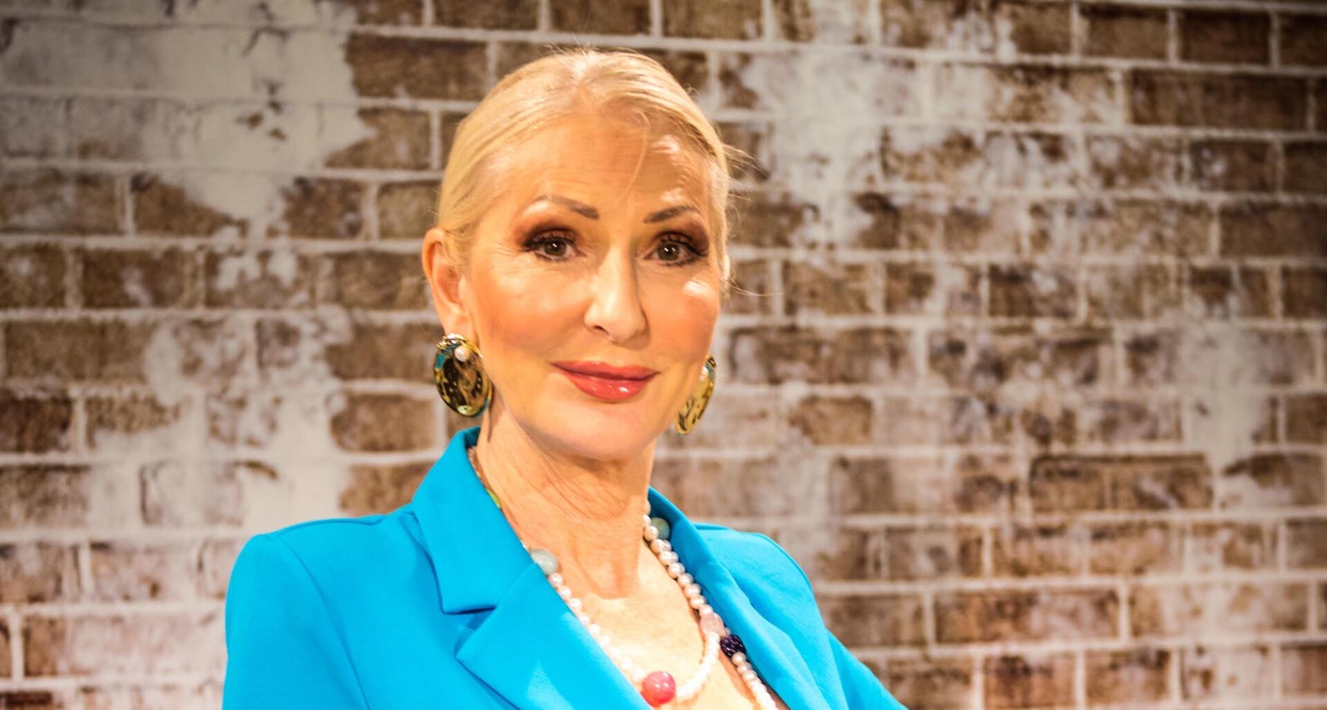 Désirée Nick trägt einen blauen Blazer, zurück gebundene Haare und steht vor einer braunen Ziegelwand.