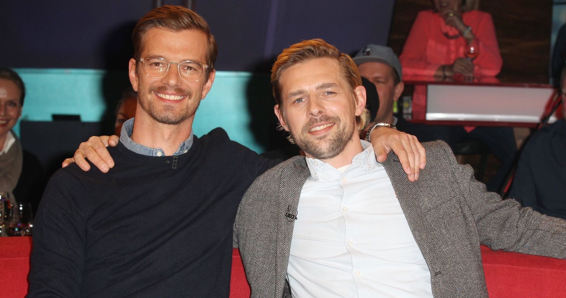 Joko Winterscheidt und Klaas Heufer Umlauf sitzen auf einer roten Bank und haben die Arme umeinander gelegt. Beide lachen in die Kamera.