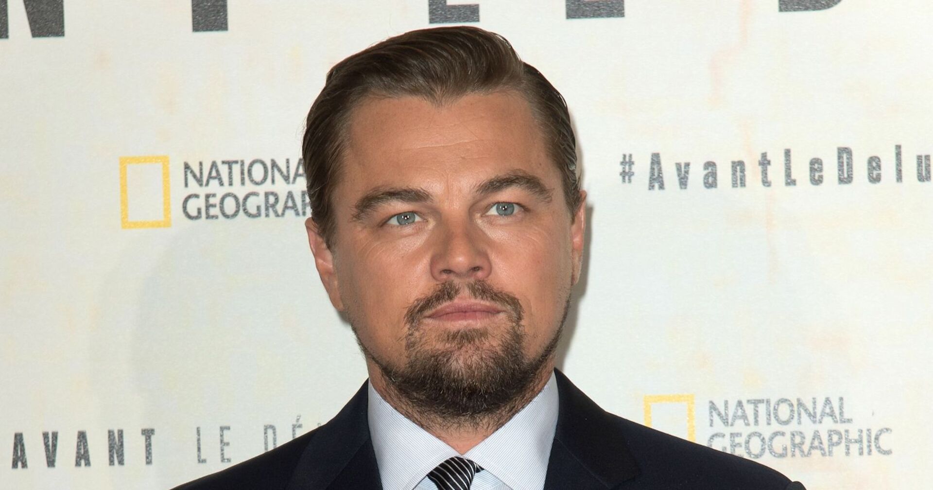 Leonardo Di Caprio schaut streng in die Ferne. Er steht vor einer National Geographic Leinwand und trägt einen schwarzen Anzug.