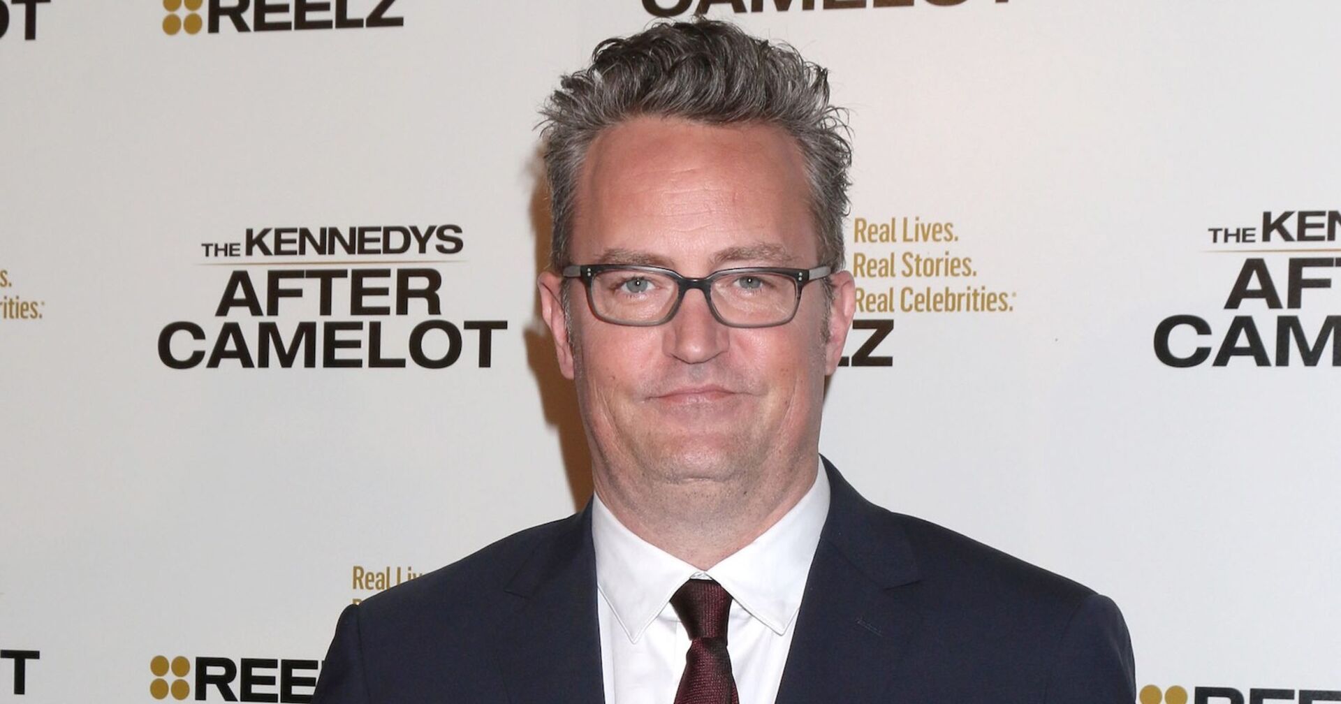 Matthew Perry trägt ein schwarzes Sakko und eine weiße Bluse und eine Brille. Er steht vor einer Fotoleinwand.