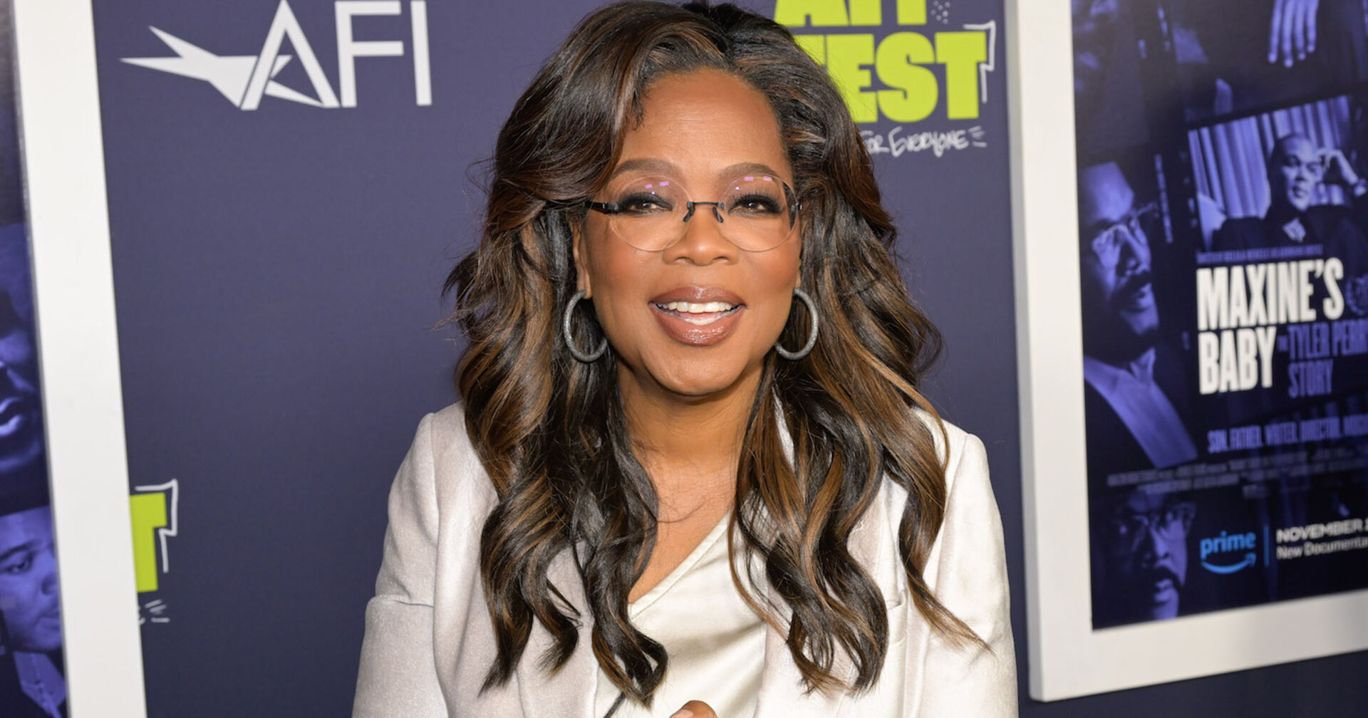 Oprah Winfrey mit einer weißen Bluse und offenen gewellten Haaren. Sie trägt große Ohrringe und eine Brille.