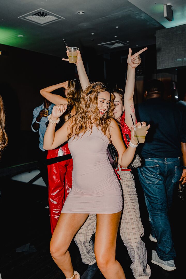 Zwei Frauen tanzen ausgelassen zu einer Party. Eine trägt ein kurzes Beiges Kleid und die andere eine Hose.