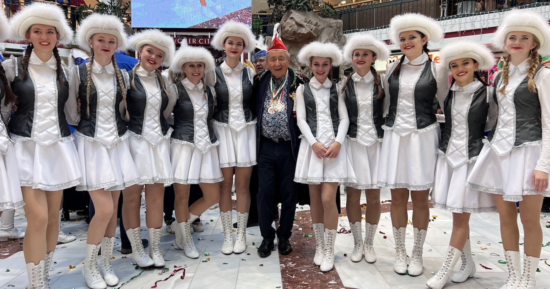 Richard Lugner trägt einen roten Hut. Er ist umgeben von Tänzerinnen mit weißen Outfits, weißen Schuhen und weißen Federhüten.