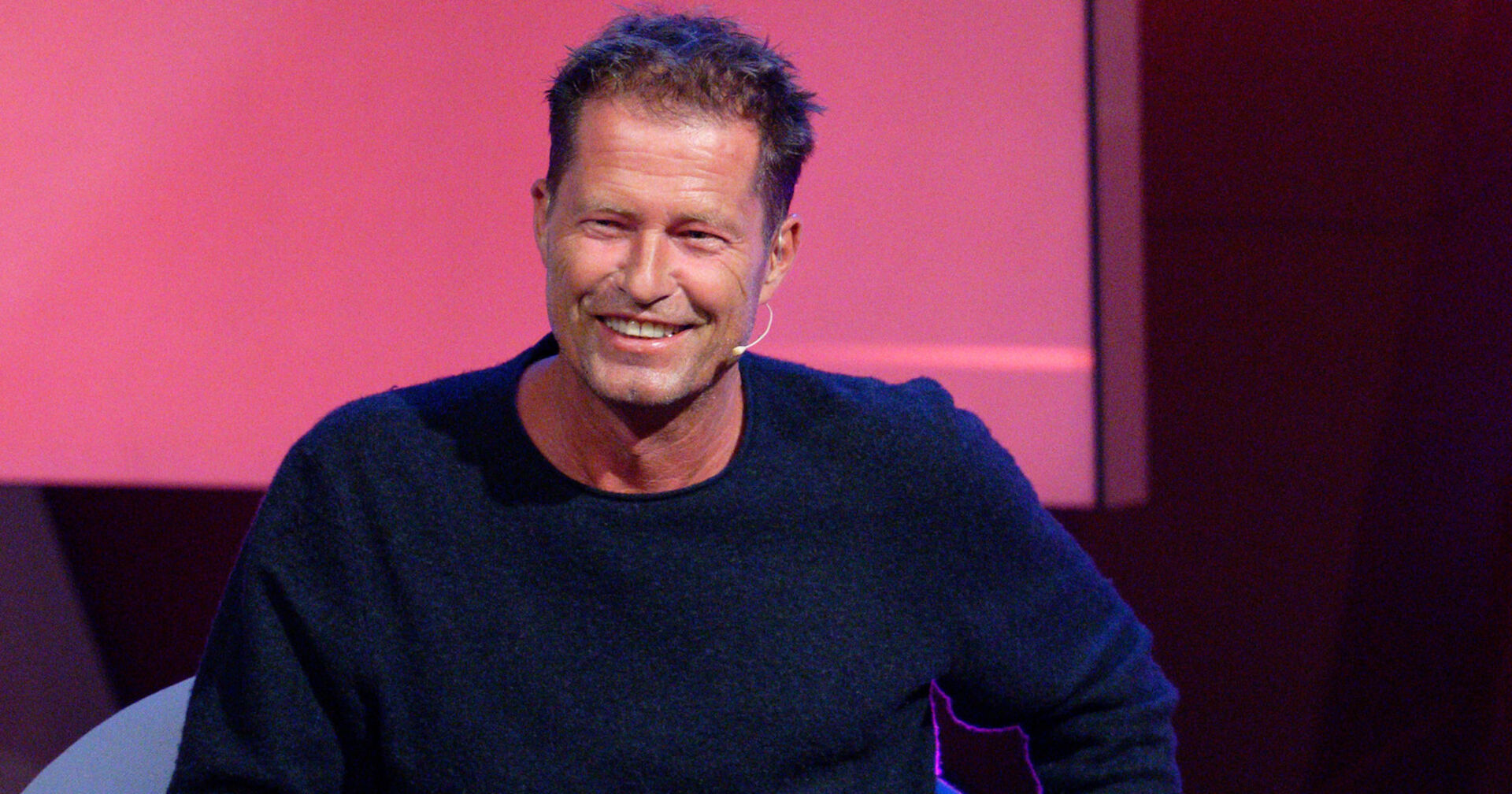 Til Schweiger mit einem dunkelblauen Pullover vor einer Rosinen Filmwand.