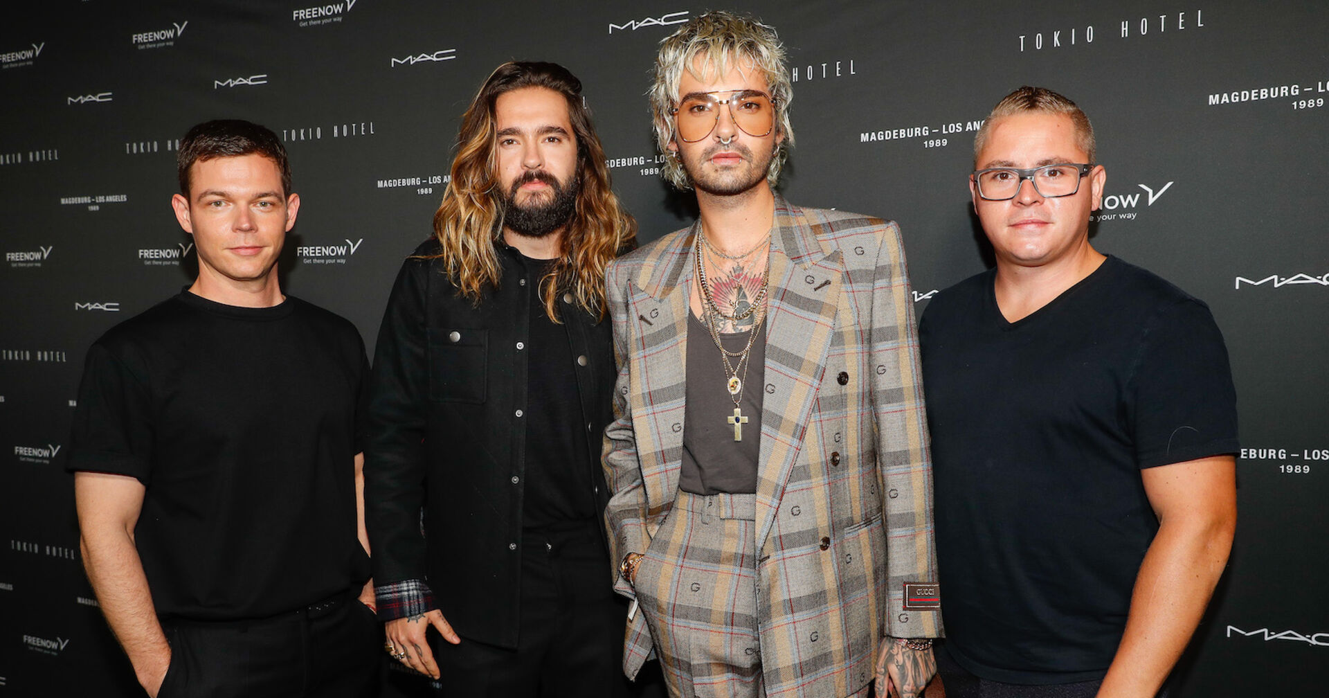 Bill Kaulitz trägt einen karierten Anzug, seine Bandmitglieder sind schwarz angezogen. Sie sehen vor einer schwarzen Fotoleinwand.