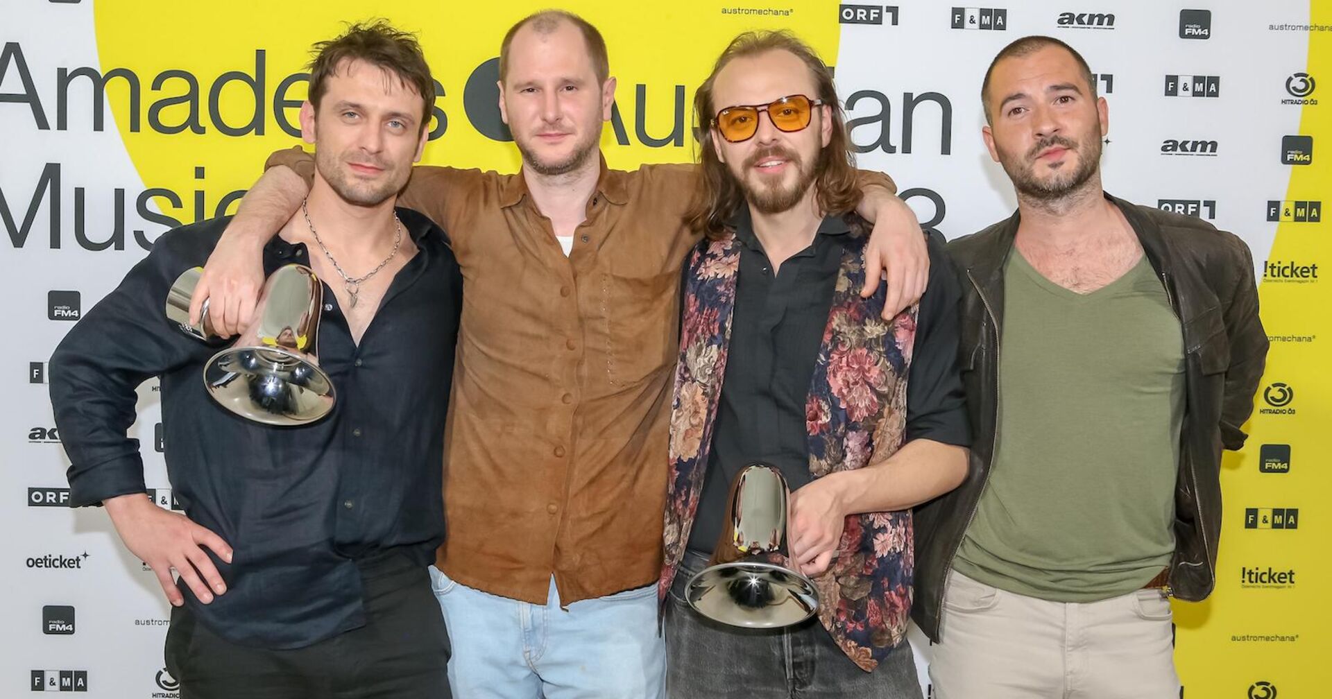 Wanda beim Austrian Musik Award. Sie tragen lässige Kleidung, halten zwei Megafone und stehen vor einer Fotowand auf der steht "Amadeus Music Award".