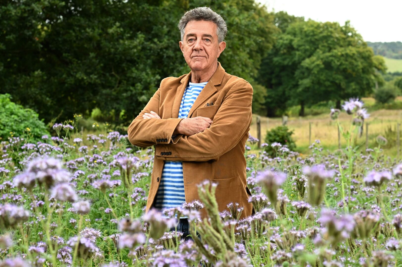 Yves Cochet, der ein braunes Samtsakko trägt und mit verschränkten Armen in einem Blumenfeld steht