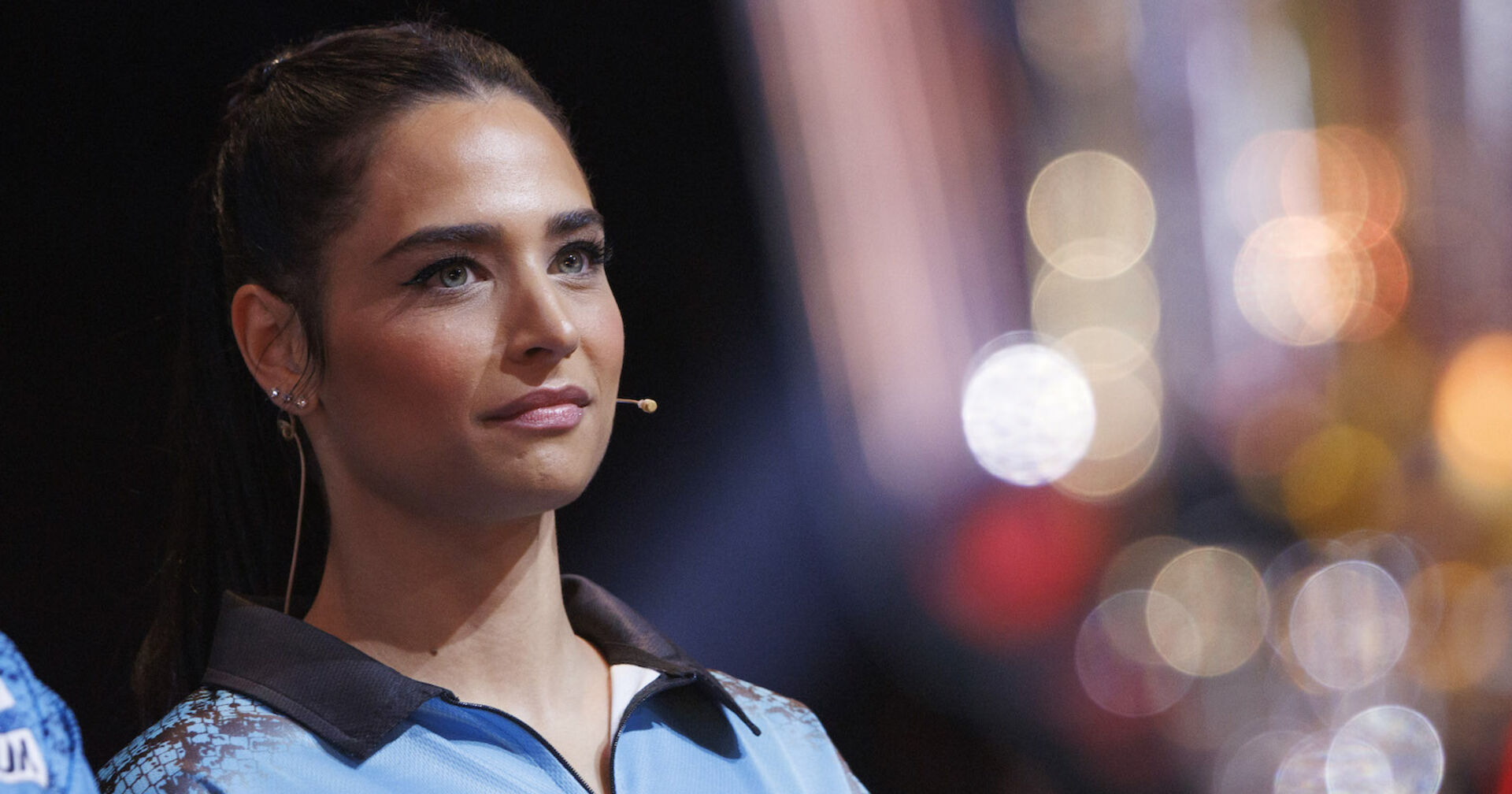 Amira Pocher in einem hellblauen Sportshirt. Sie sieht in die Ferne und hat ein Mikrofon an ihrer Wange montiert.