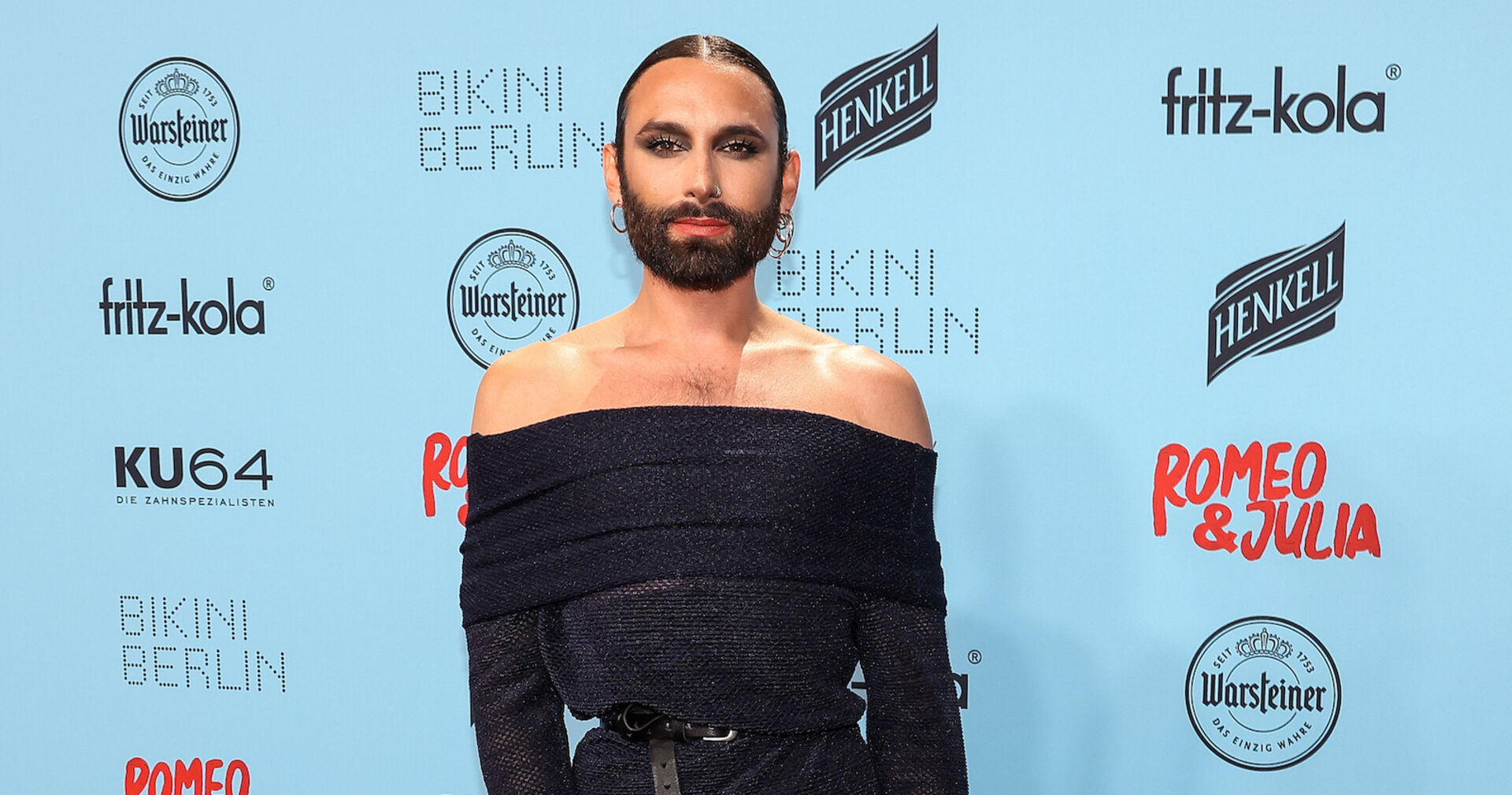 Conchita Wurst mit einem engen schwarzen Kleid und gegelten Haaren vor einer hellblauen Fotowand.