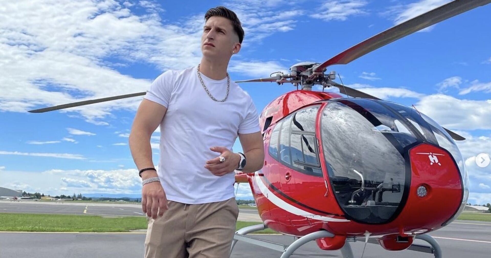 Satans Bratan mit einem weißen T-Shirt vor einem roten Helikopter.