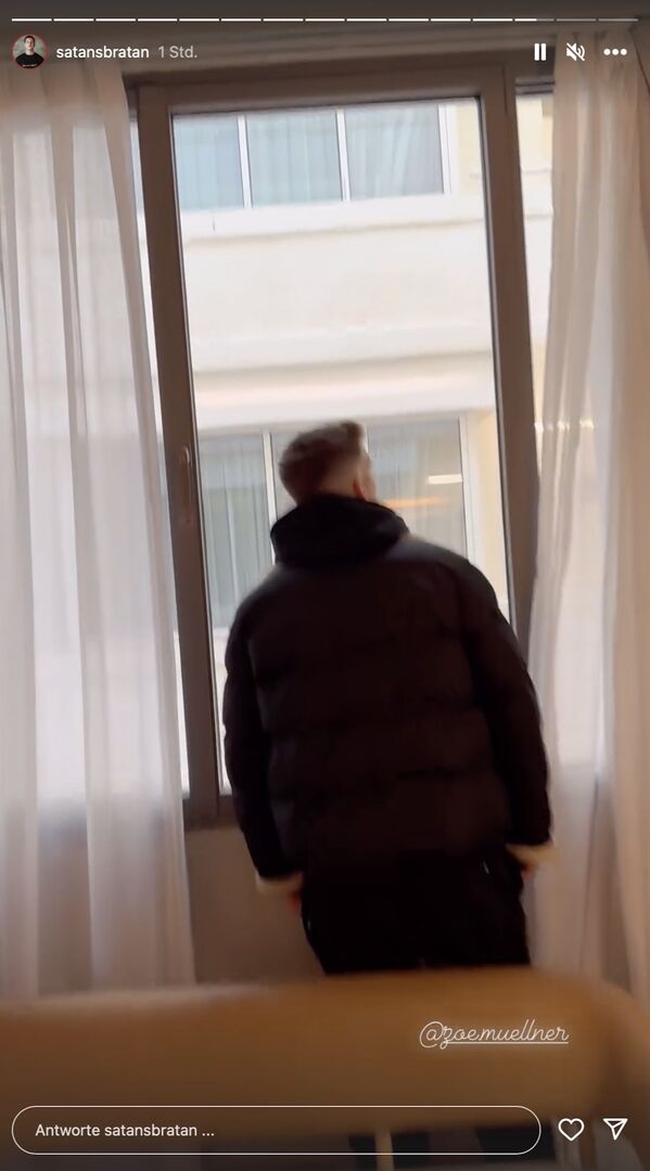 Satans Bratan vor dem Ausblick in den Innenhof in einem Hotelzimmer. Er trägt einen schwarzen Hoodie.