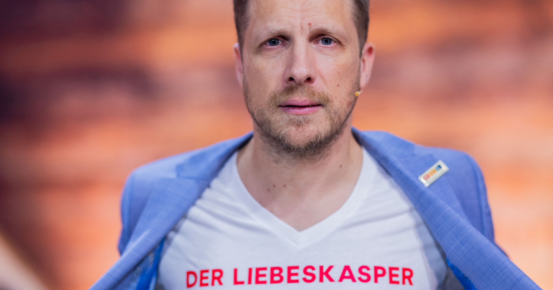 Oliver Pocher mit einem weißen T-Shirt auf dem steht "Liebeskasper" und einem hellblauen Sakko.