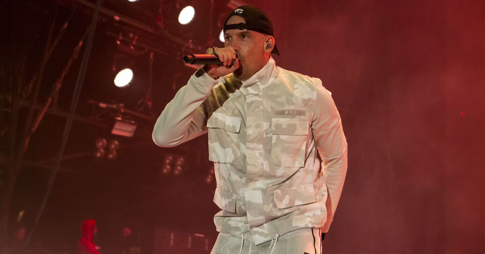 Raf Camora im weißen Outfit mit Mikrofon und Kappe auf der Bühne.