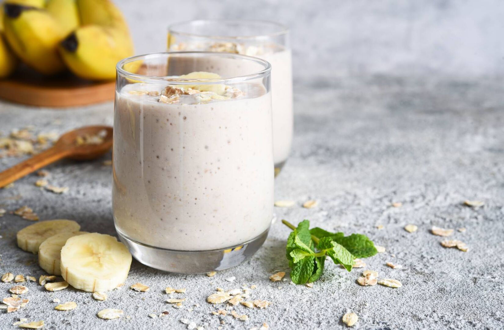 Zwei Gläser, gefüllt mit einem hellen Smoothie auf einer Steinplatte. Zur Dekoration liegen auf der Steinplatte 3 aufgeschnittene Bananenscheiben und ein kleiner Zweig mit Minzblättern.