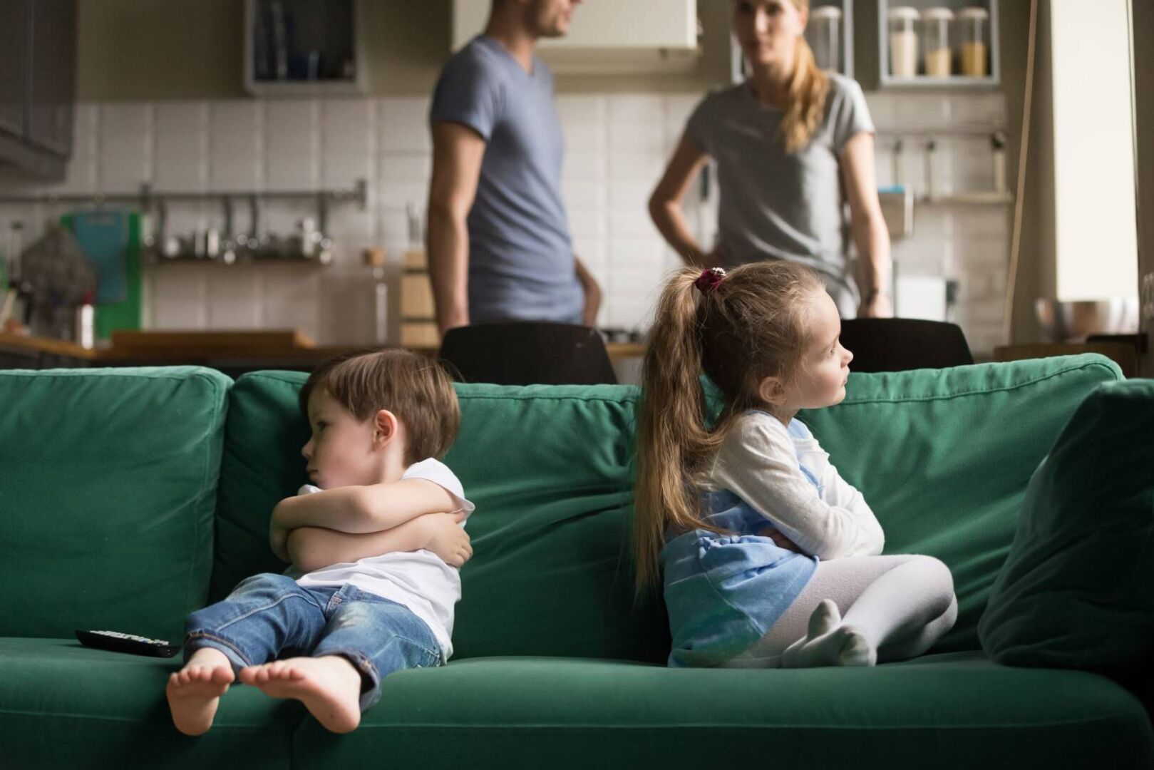 Zwei Kinder sitzen mit verschränkten Armen Rücken an Rücken auf dem Sofa.