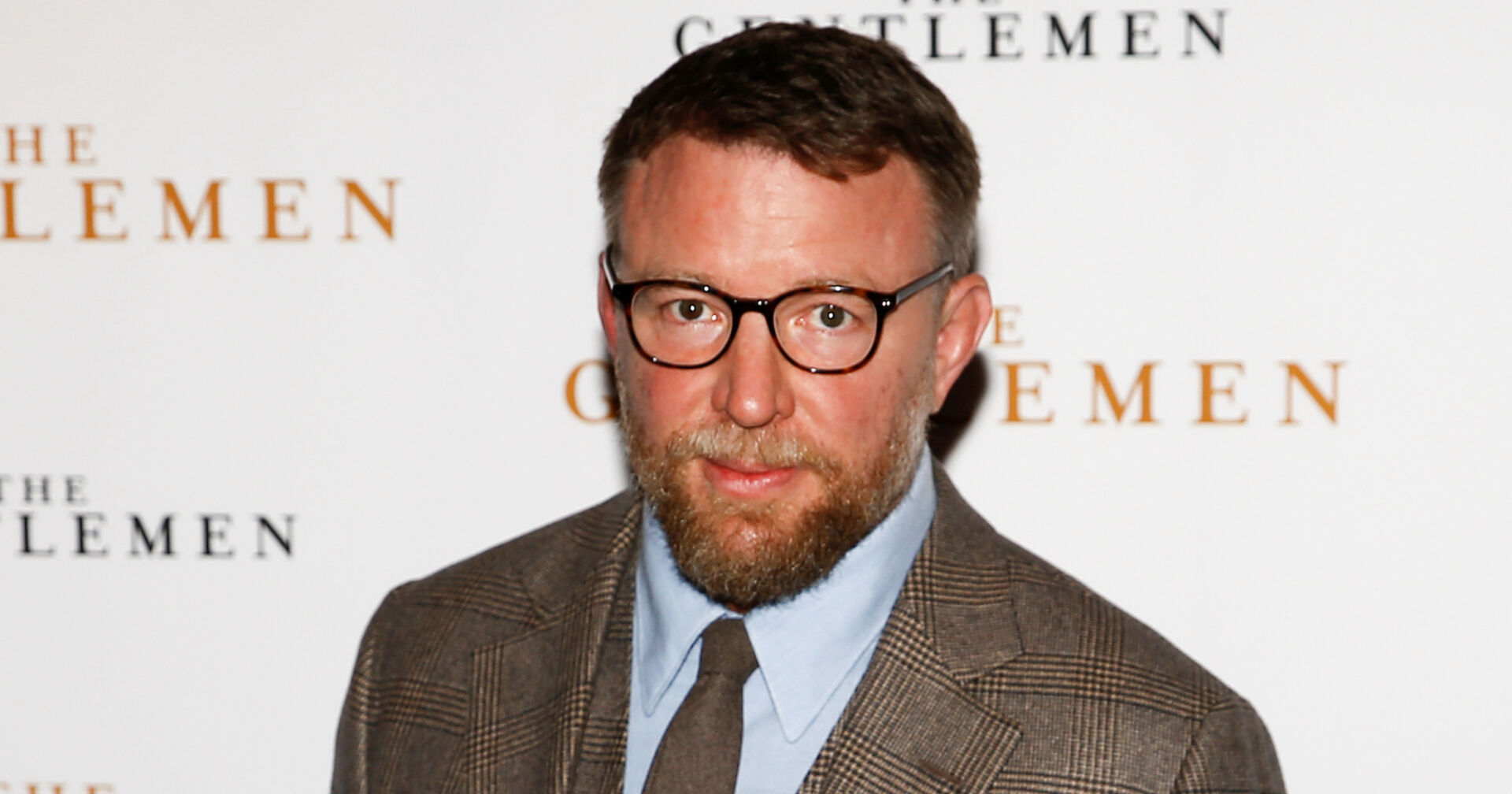 Guy Ritchie vor einer Fotoleinwand mit einem beigen Sakko.