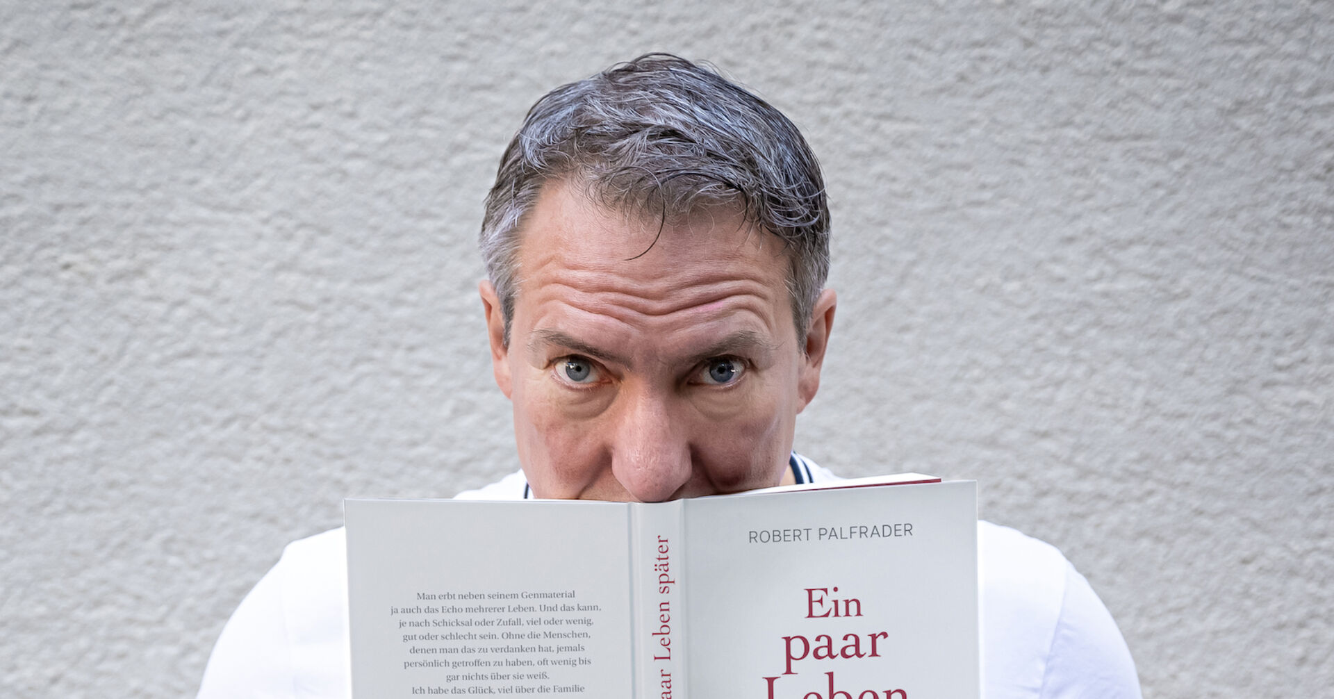 Robert Palfrader hält sein Buch "Ein paar Leben später" und schaut über den Rand hinweg.